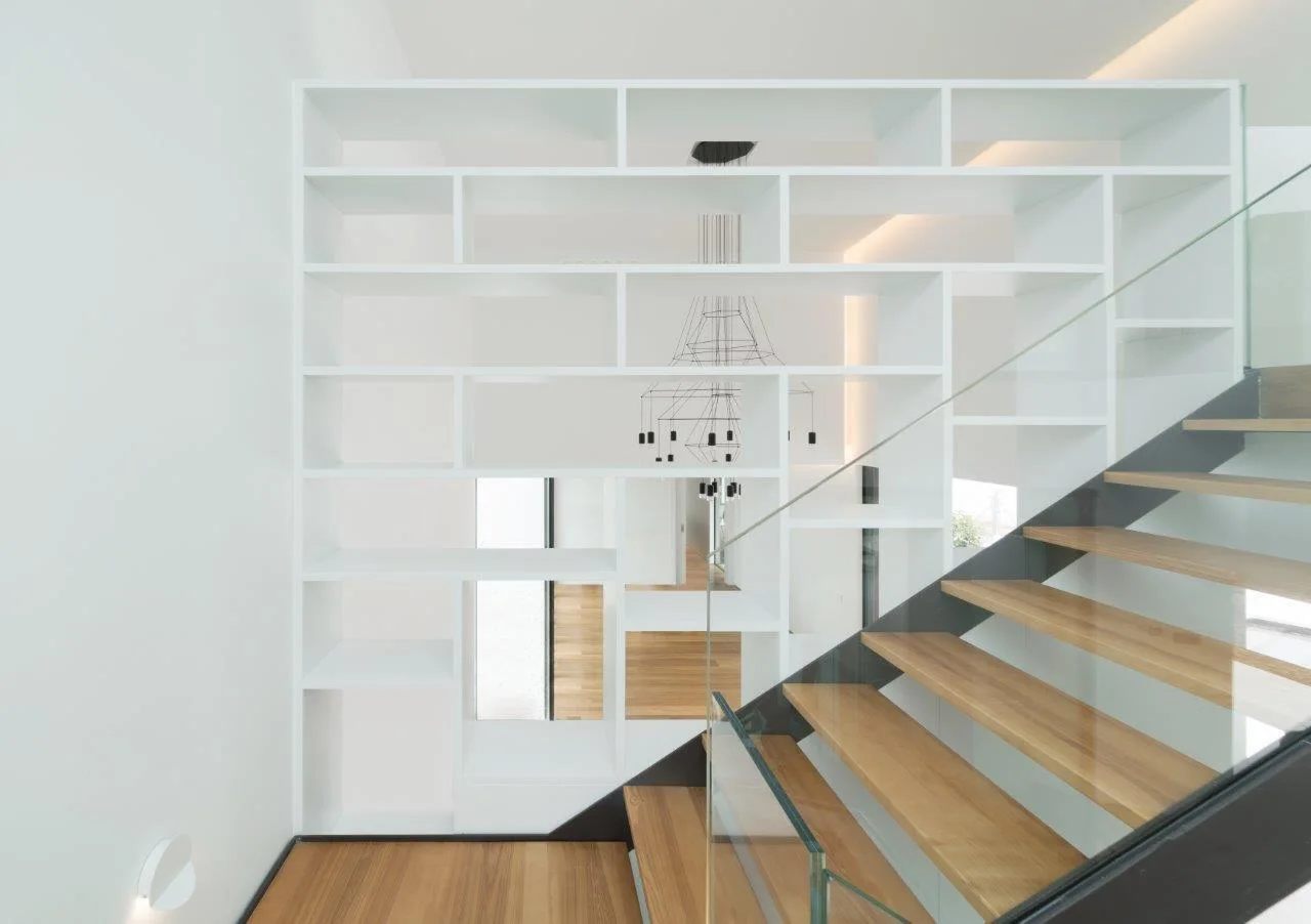 A modern wooden staircase with a glass railing stands in front of a floor-to-ceiling white open shelving unit.