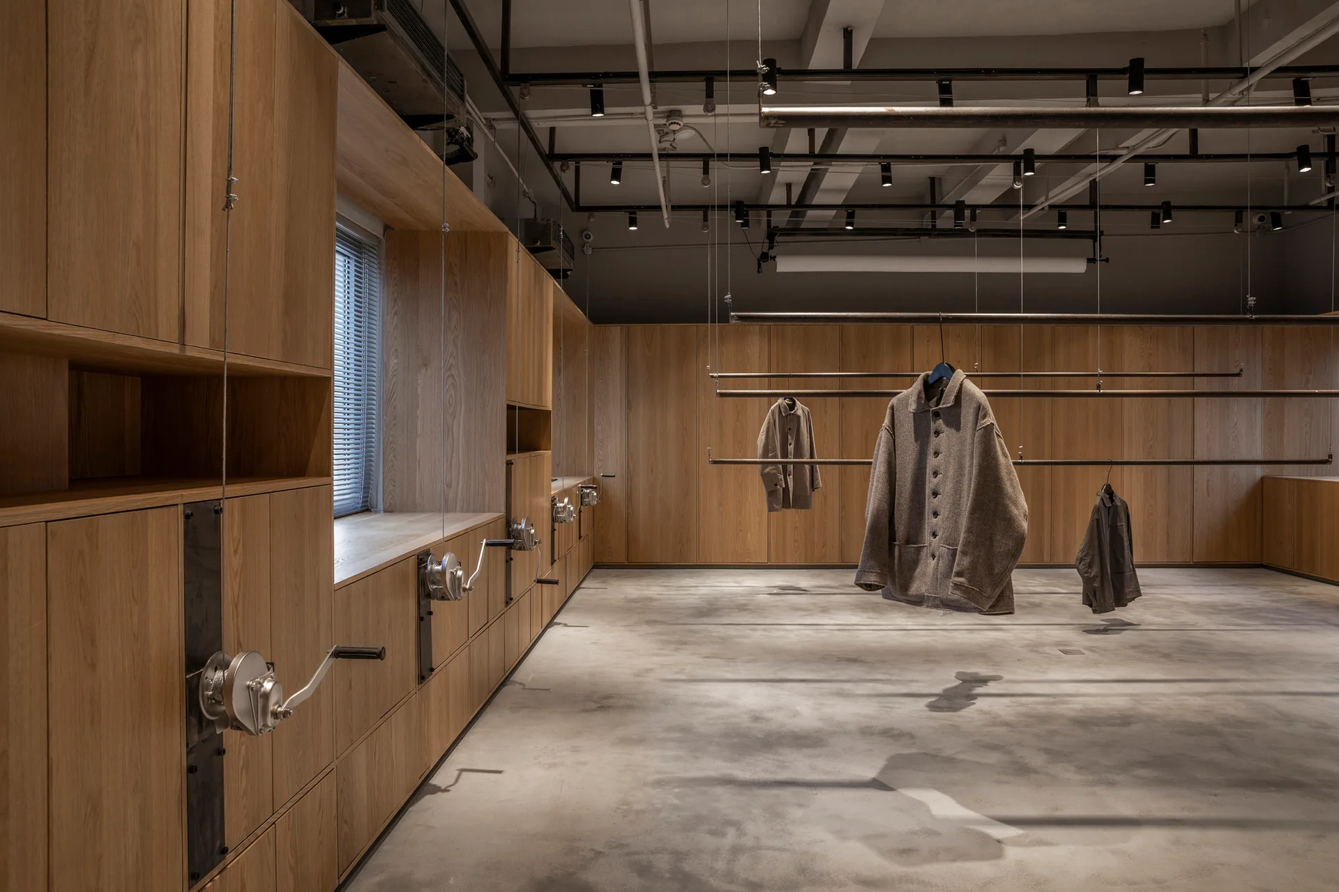 An industrial retail interior features wood-paneled wall cabinets, concrete flooring, and metal clothing racks suspended from an exposed ceiling.