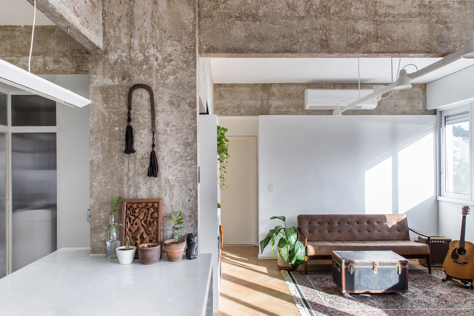 An industrial-style living room features an exposed concrete column, a brown leather sofa, and a vintage trunk coffee table on a patterned rug.