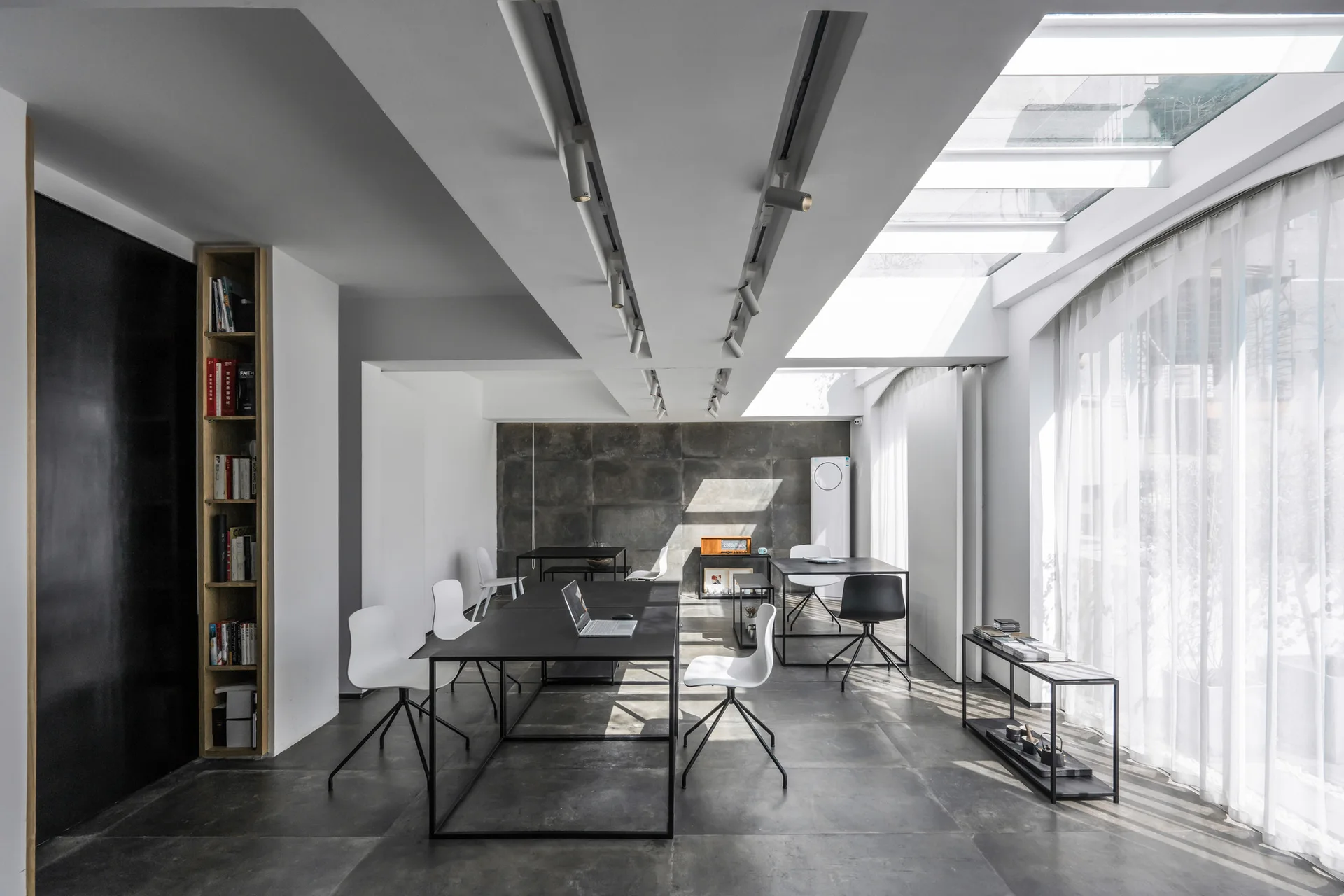 A minimalist office with black metal desks, white chairs, grey tiled floors, and a bright glass skylight roof next to sheer white curtains.