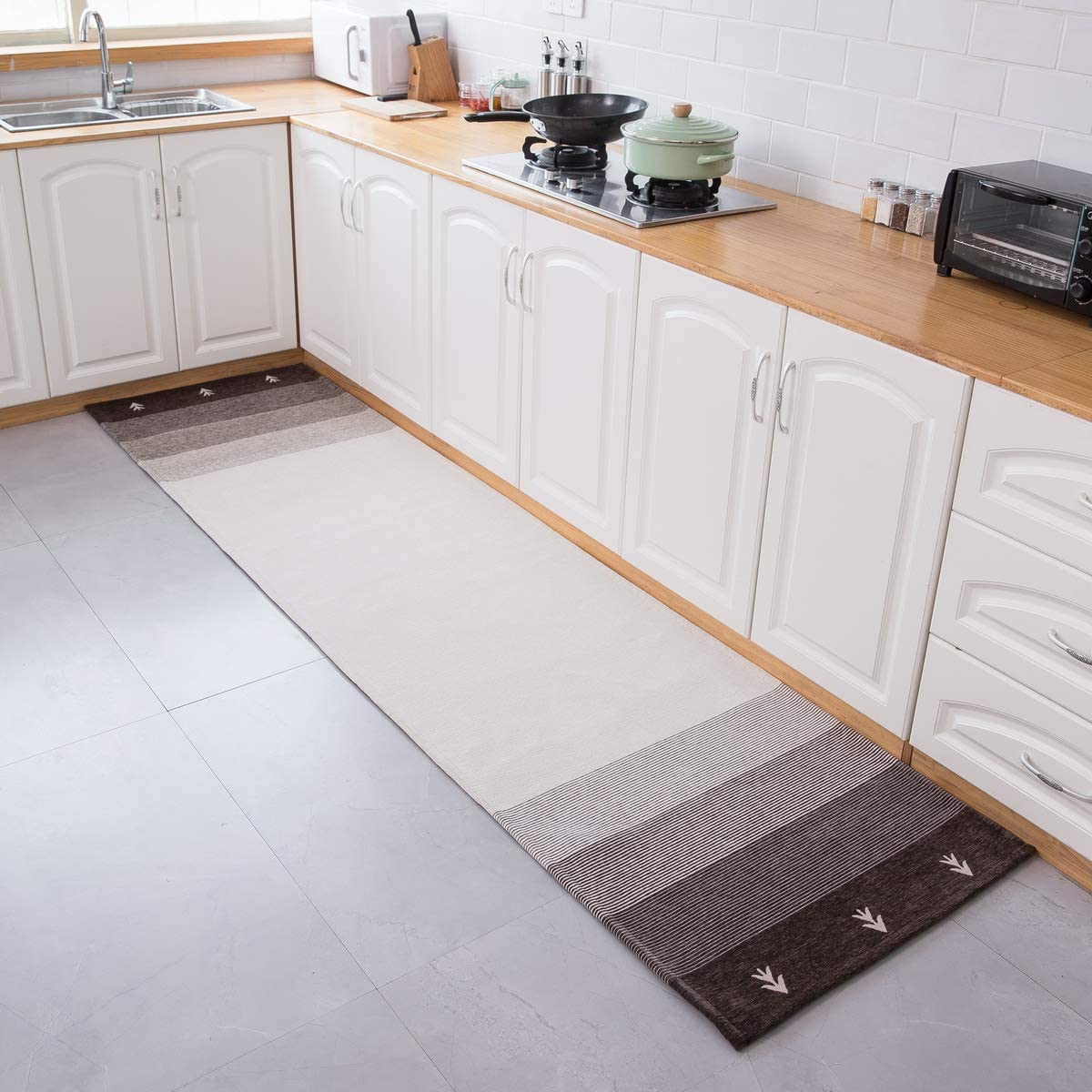 A kitchen featuring white shaker cabinets, a light wood countertop, a white subway tile backsplash, and a long striped runner rug on grey floor tiles.