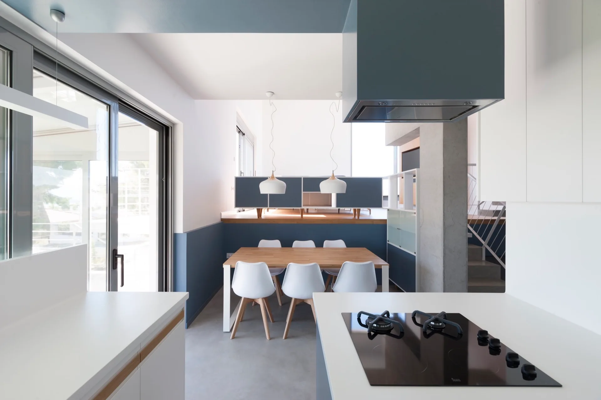 A modern split-level kitchen features a foreground stovetop, a wooden dining table with white chairs, and blue painted walls.
