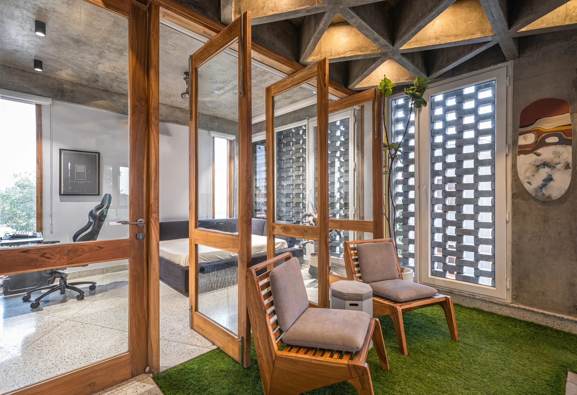Two wooden lounge chairs sit on artificial grass flooring beneath a geometric concrete ceiling, separated from interior rooms by wood-framed glass doors.