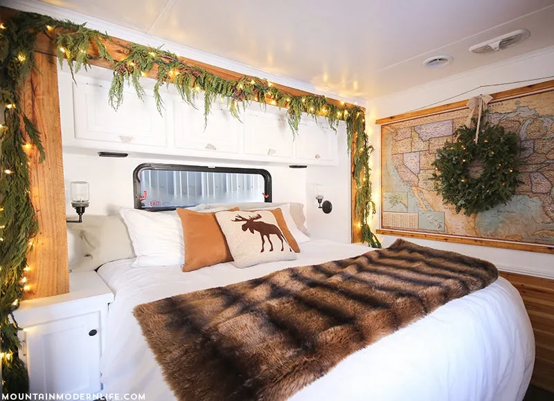 A small camper bedroom features a bed with a faux fur throw, white overhead cabinets, and a rustic wood frame decorated with holiday garland and lights.