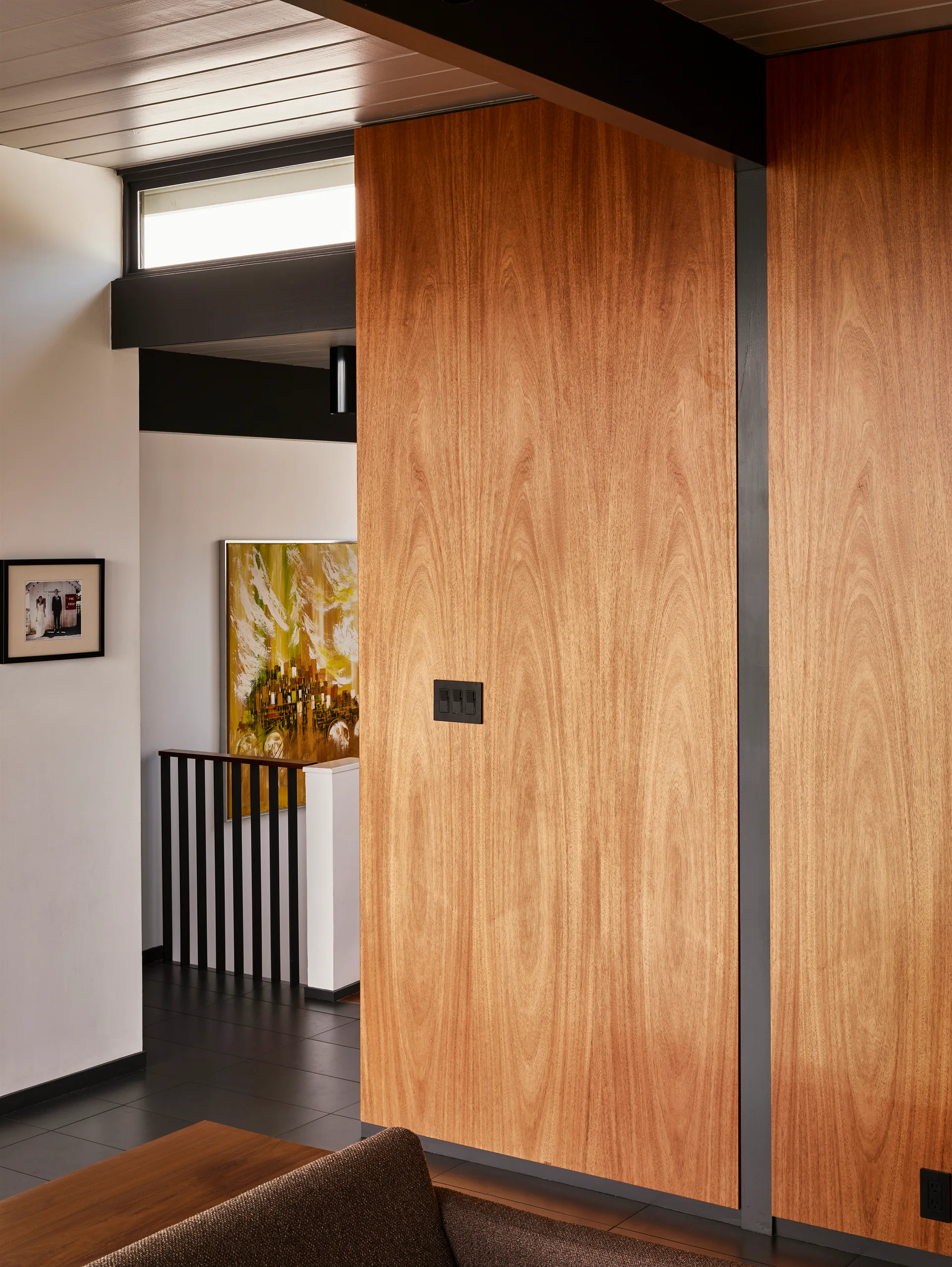 A residential hallway features a warm wood-paneled wall contrasted by a dark ceiling beam, a transom window, and a black stair railing.