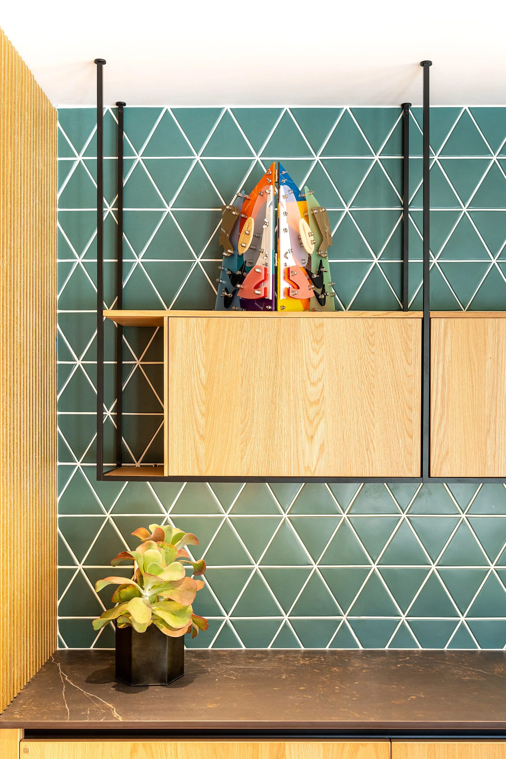 A kitchen wall featuring green triangular tiles with white grout, suspended wood cabinets, a vertical wood slat wall, and a dark countertop.