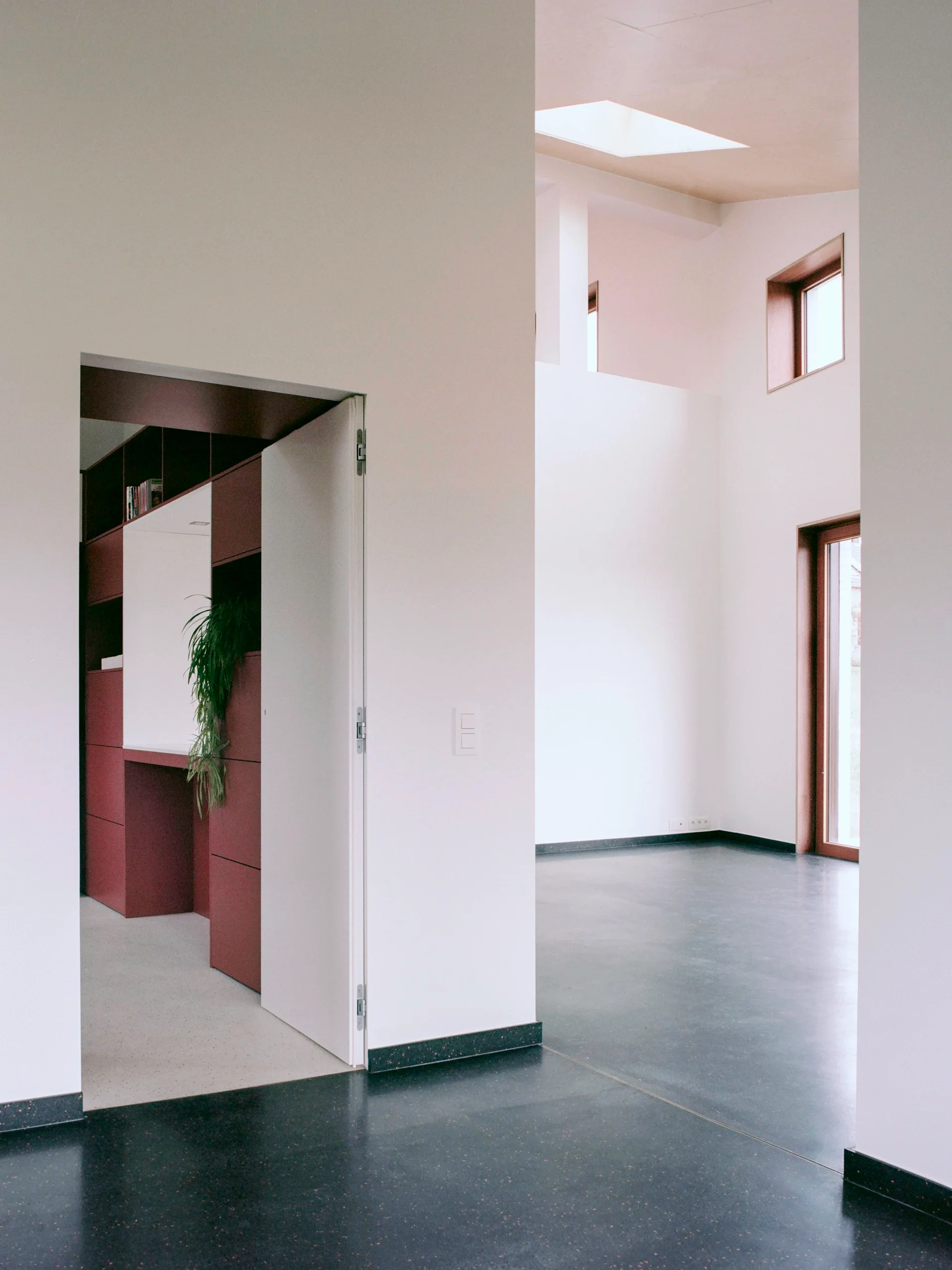 A modernist home interior featuring a dark grey floor, white walls, built-in maroon cabinetry, and a high-ceilinged room with a skylight.