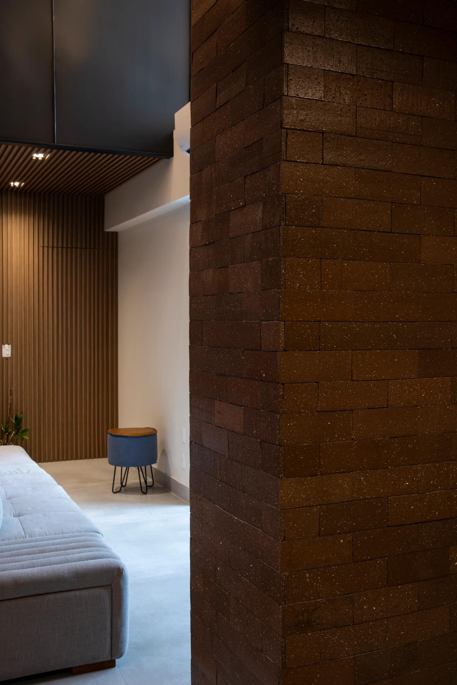 A bedroom featuring a large textured brown brick column in the foreground, a grey tufted bed, a blue stool, and a vertical wood slatted wall.