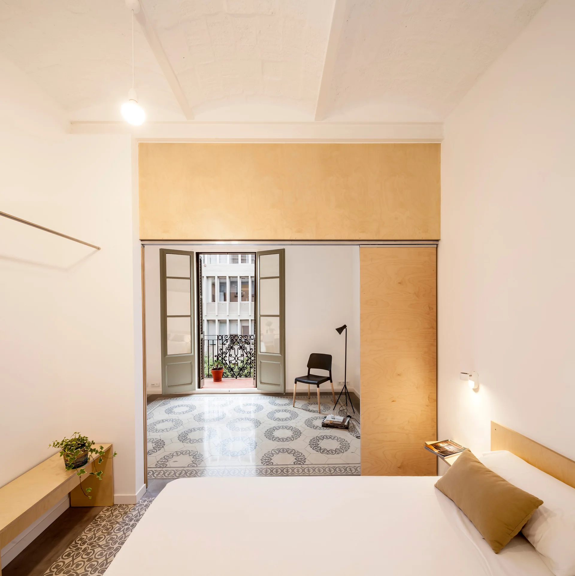 A minimalist bedroom with a large plywood sliding partition opening to an adjacent room with patterned floor tiles and French doors.