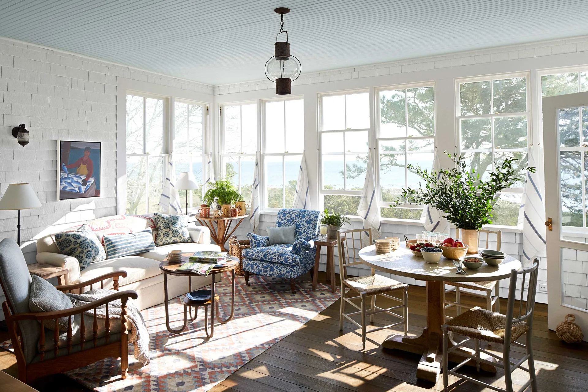 A bright coastal sunroom featuring white shingle walls, a light blue ceiling, a cream sofa, and a wooden dining table by large windows.