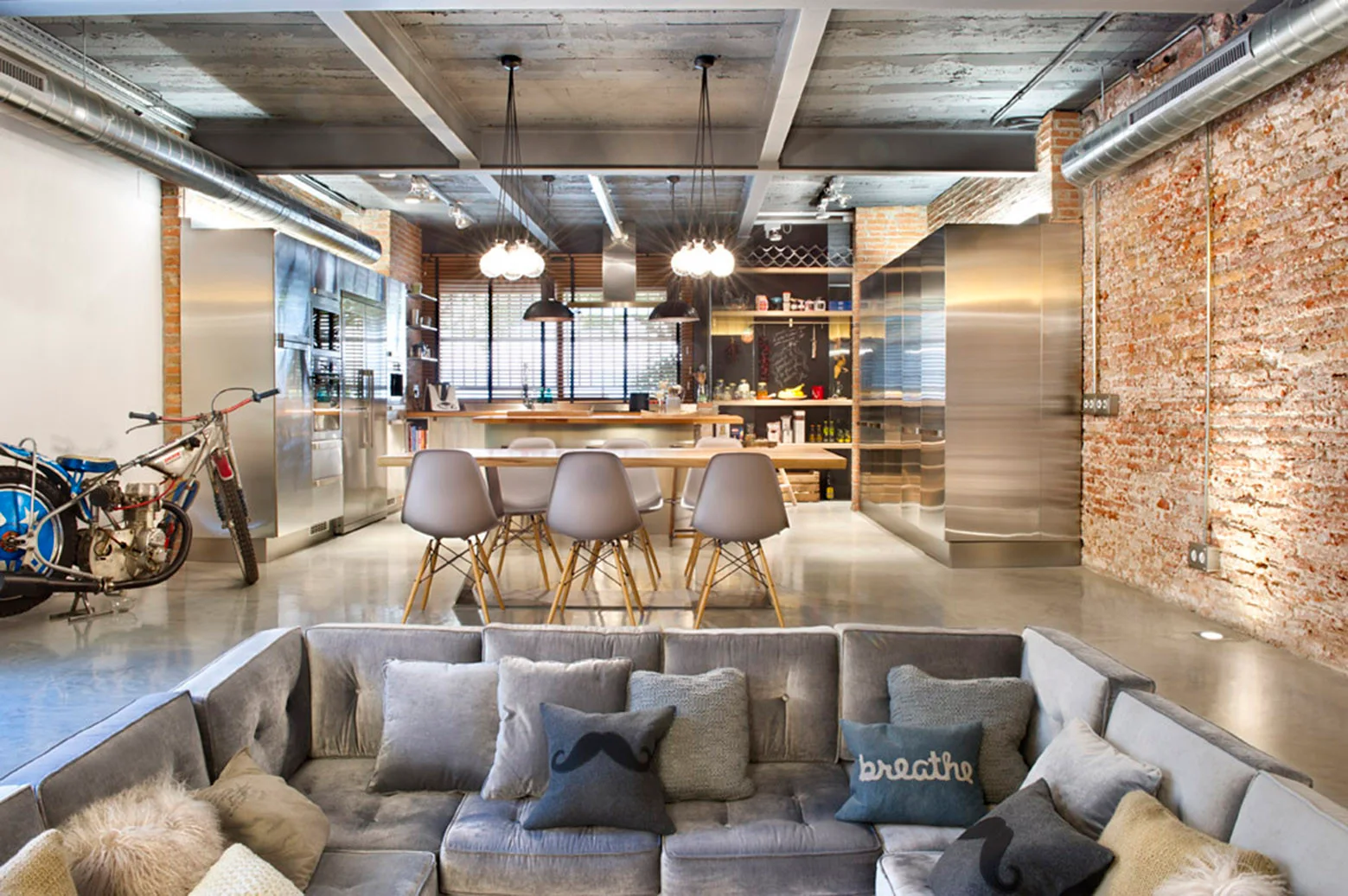 An open-concept industrial loft features a grey tufted sectional sofa, a brick accent wall, stainless steel cabinets, and concrete floors.