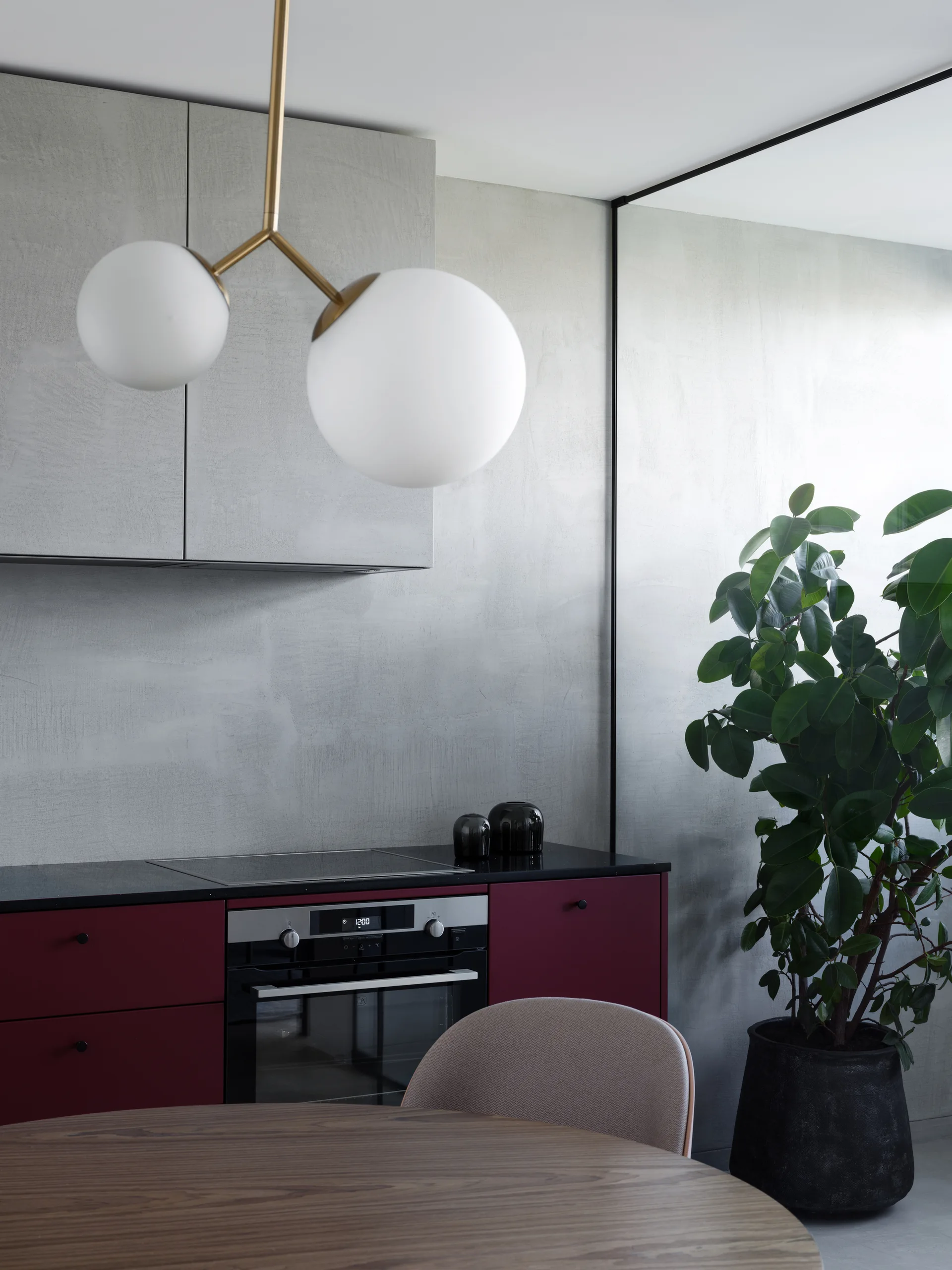 A modern kitchen featuring deep red lower cabinets, a textured grey wall, and a brass dual globe pendant light hanging above a wooden dining table.
