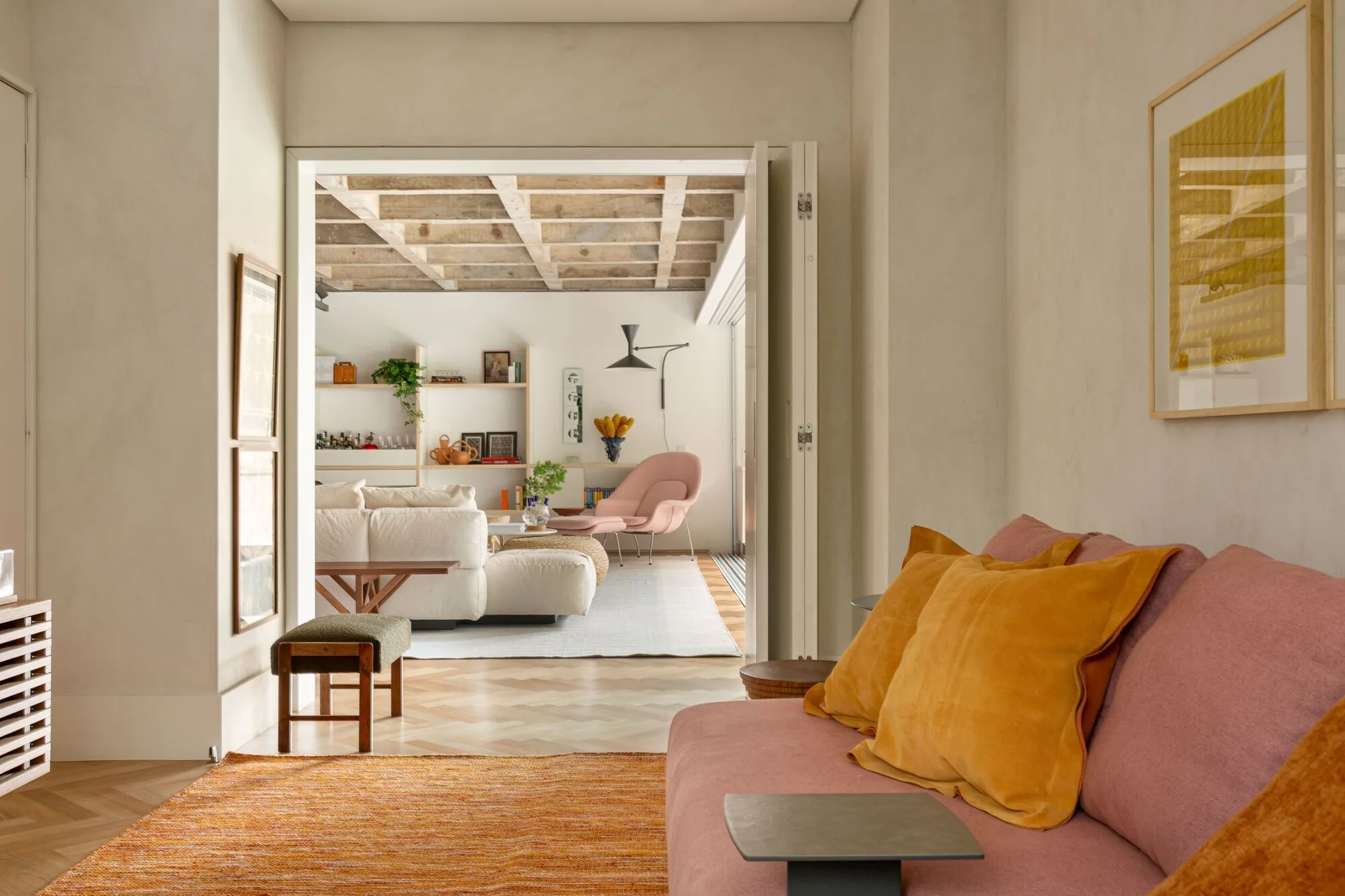 A view through an open doorway showing two connected living spaces with a pink sofa, herringbone wood floors, and an exposed wood beam ceiling.