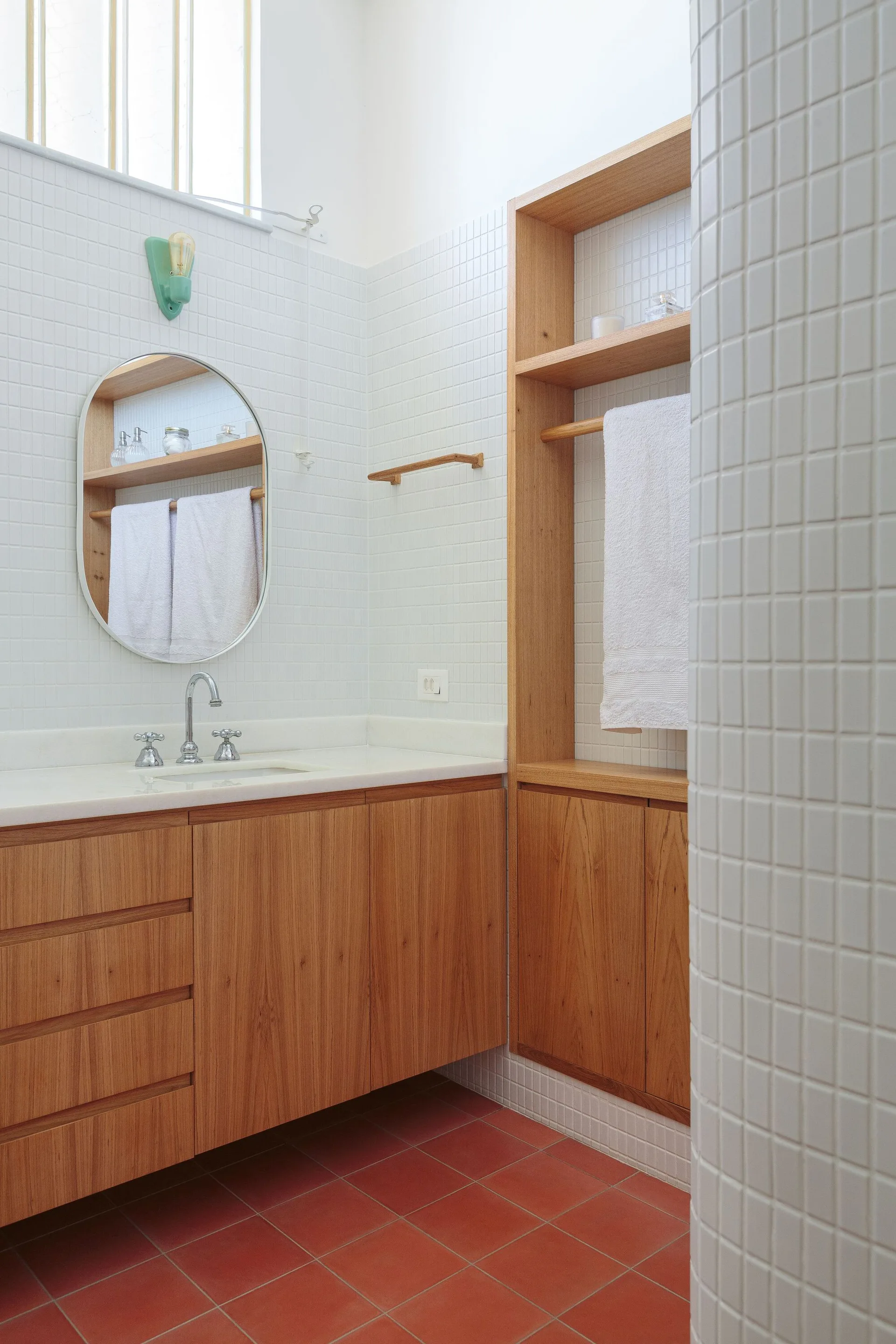 A modern bathroom features a wood vanity, an oval mirror, and integrated wood shelving set against white square wall tiles and red floor tiles.