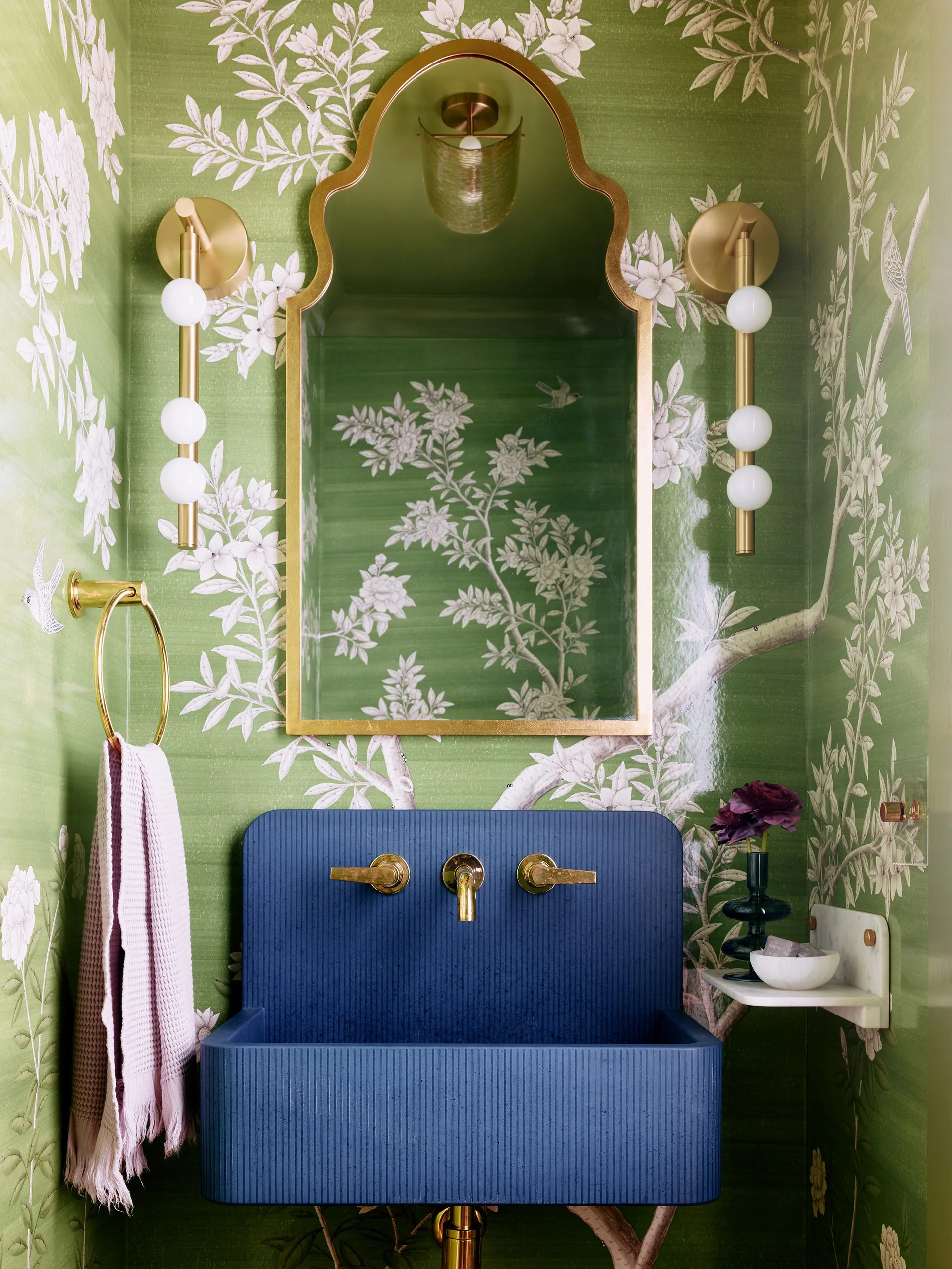 A compact powder room featuring green botanical wallpaper, a wall-mounted blue fluted sink with brass hardware, and an arched gold mirror.