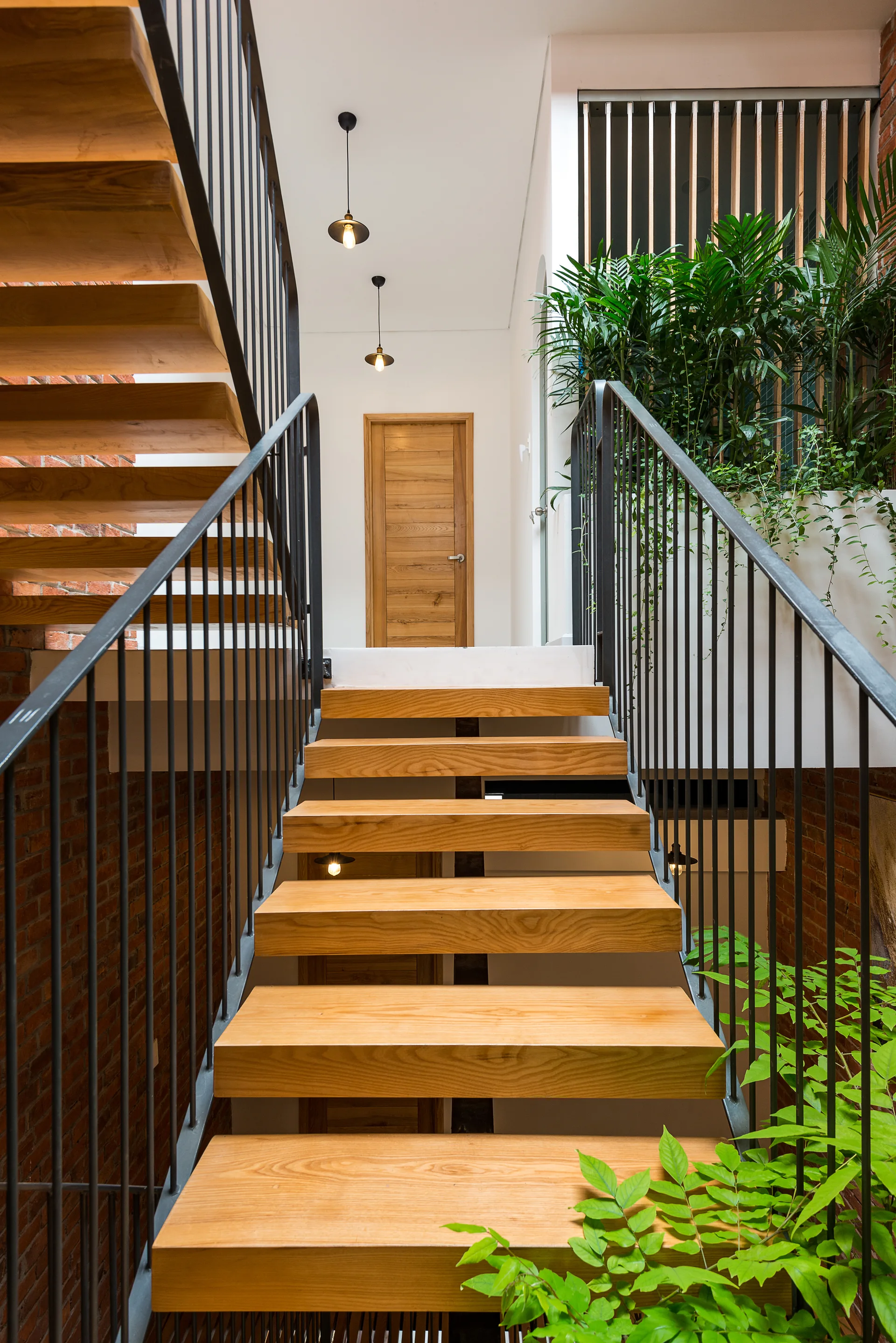 A wooden staircase with thin black metal railings rises past an exposed brick wall toward a matching wood door and green plants.