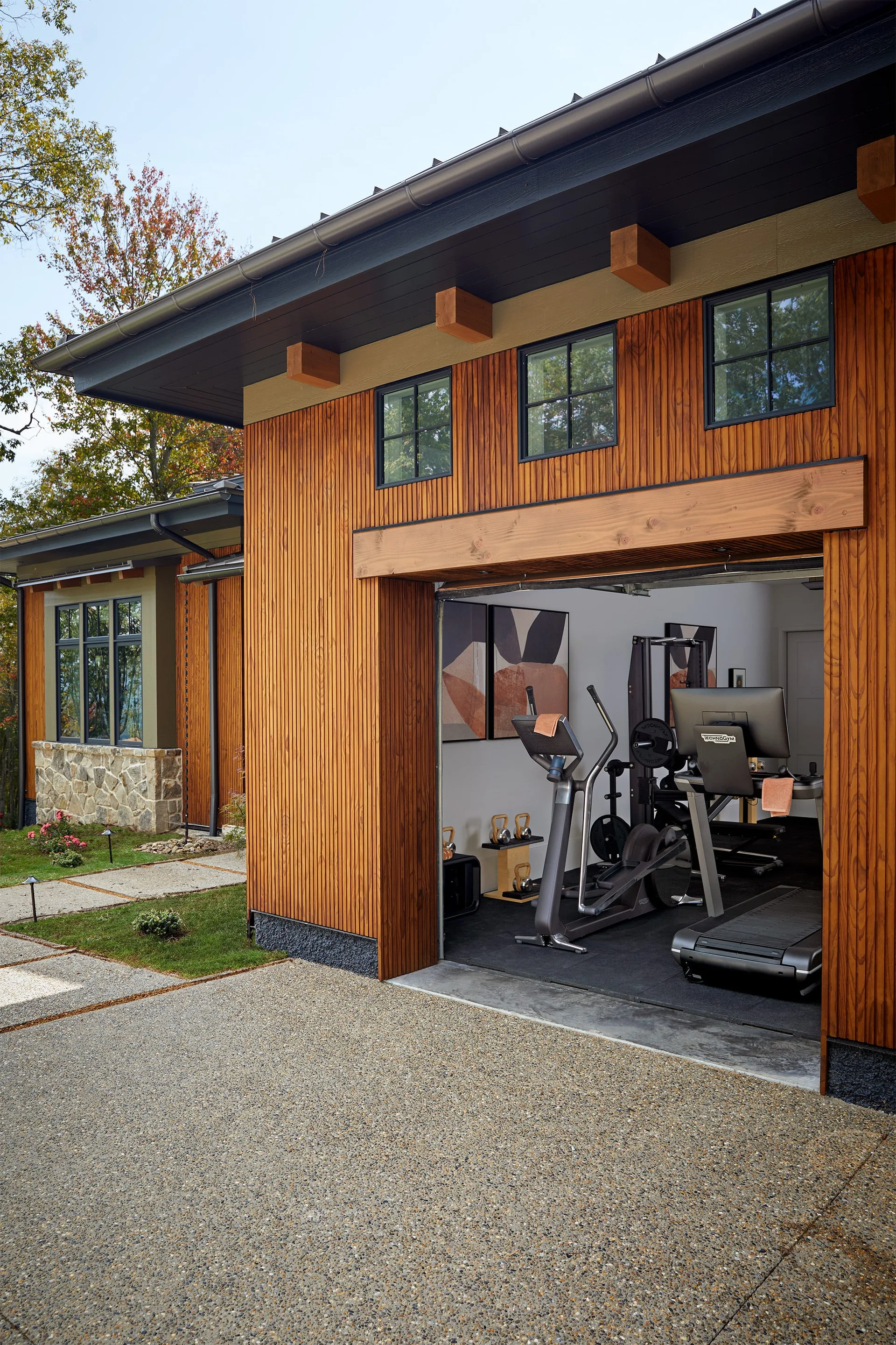 A converted garage home gym with a treadmill and elliptical trainer features an open door framed by warm vertical wood siding.