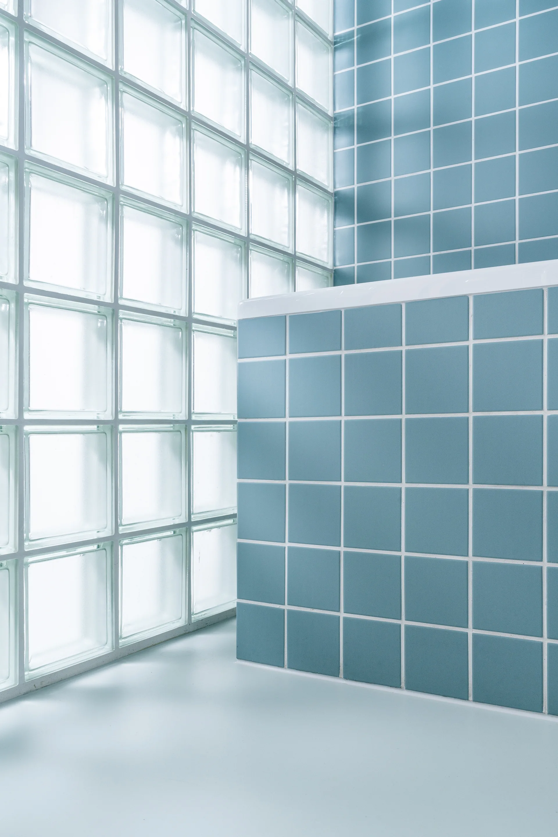 A corner of a bathroom with matte blue square tiled walls and a large, translucent glass block window letting in soft light.