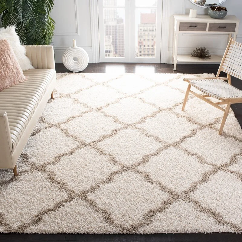 A light-filled living room features a plush cream shag rug with a brown diamond pattern, flanked by a channel-tufted sofa and a woven accent chair.