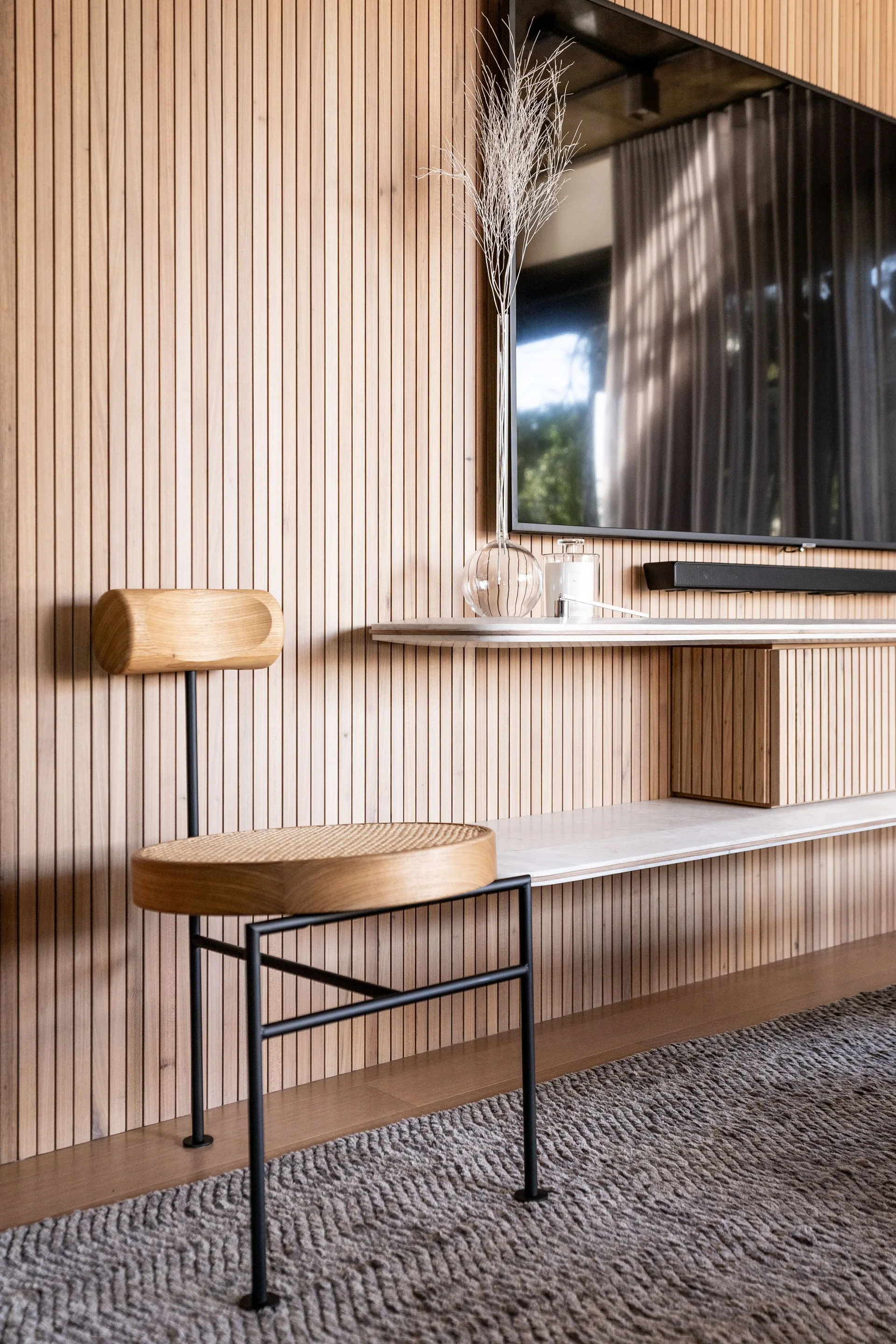 A modern living room features a vertical wood-slat wall paneling, a wall-mounted television, a stone floating shelf, and a wooden chair with a cane seat.