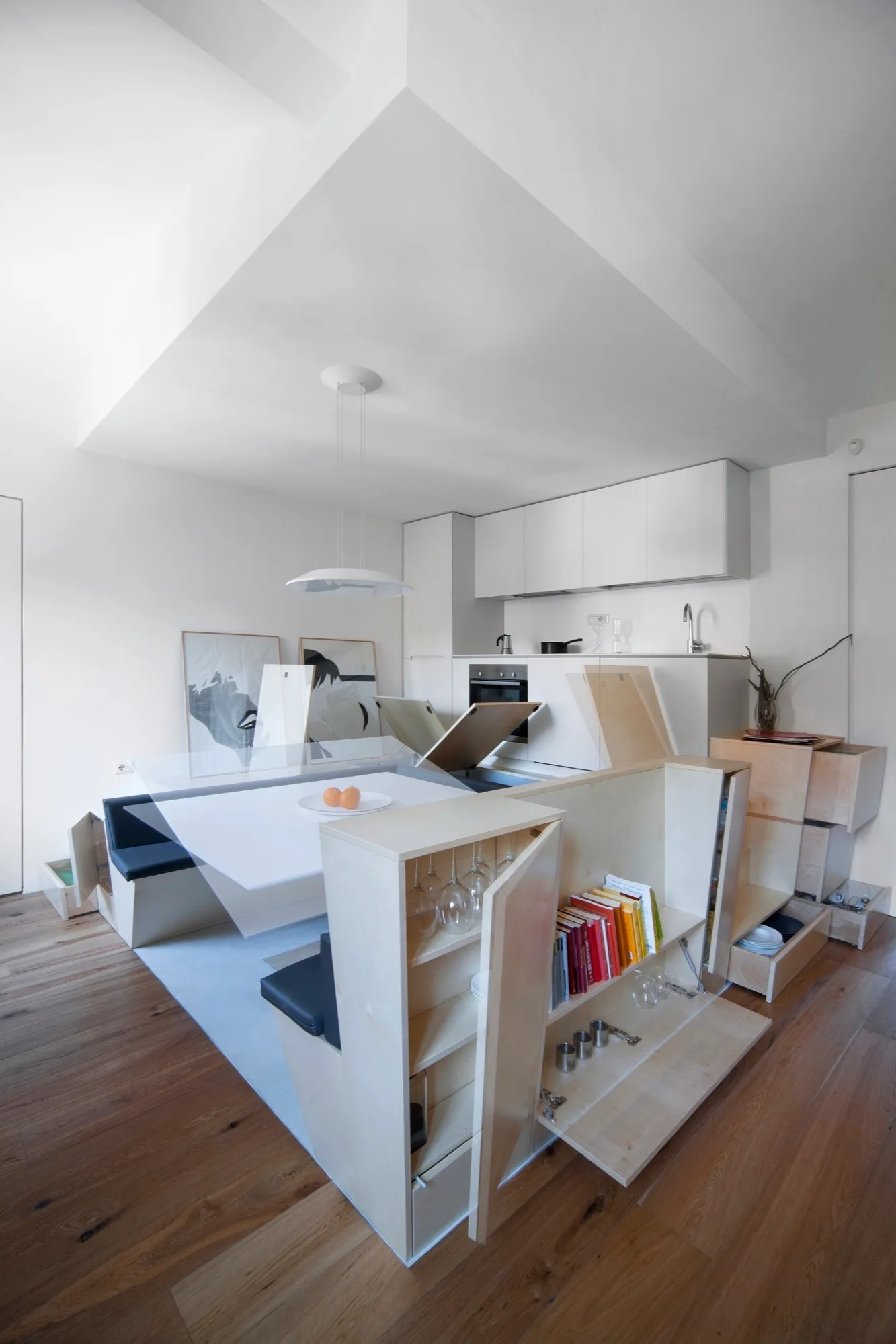 A modular plywood cabinet system with built-in bench seating and a dining table sits in a minimalist white kitchen with wood flooring.