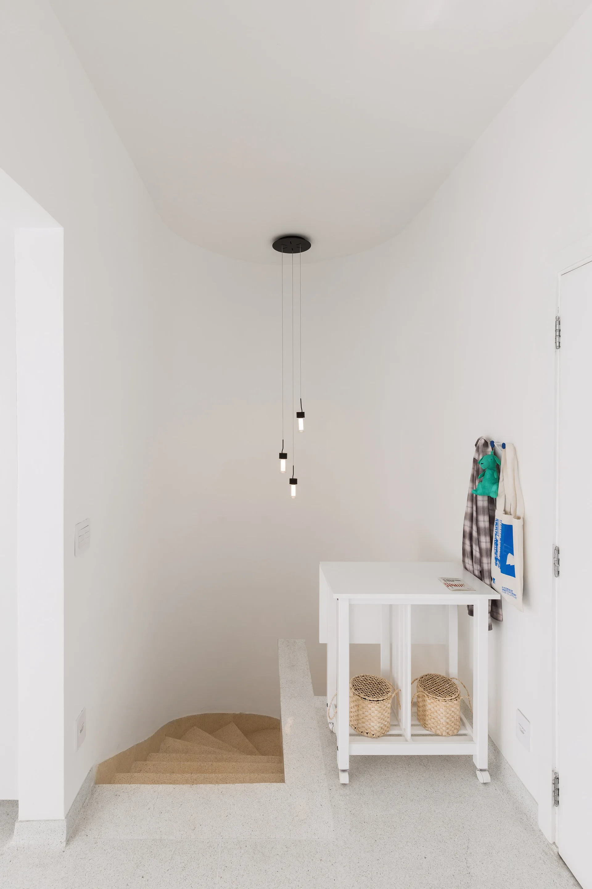 A white stairwell landing with a curved staircase, a rolling console table with two wicker baskets, and a black three-light pendant fixture.