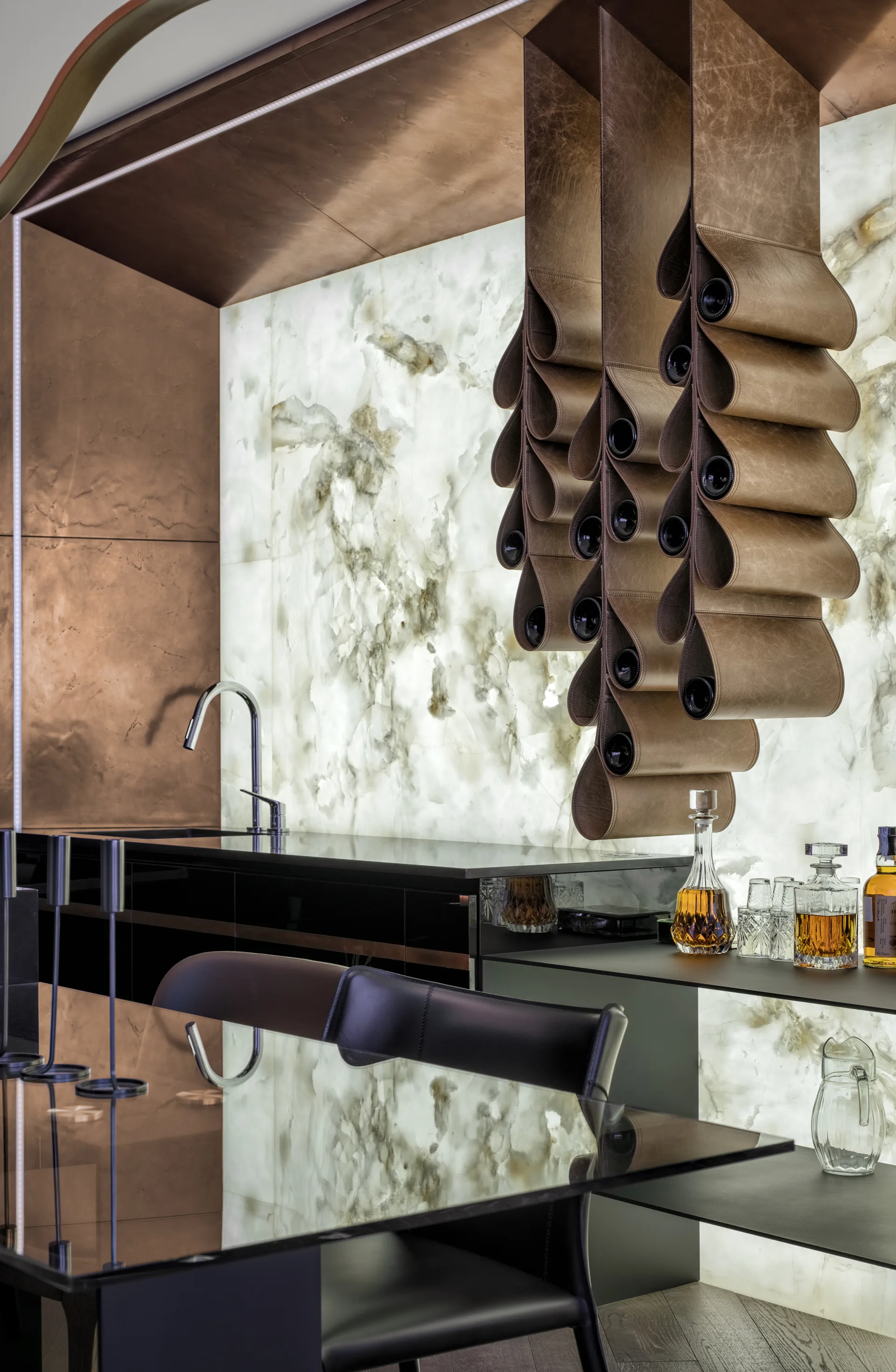 A modern home bar features a backlit stone wall, a copper-paneled alcove, and suspended leather wine storage above a black counter.