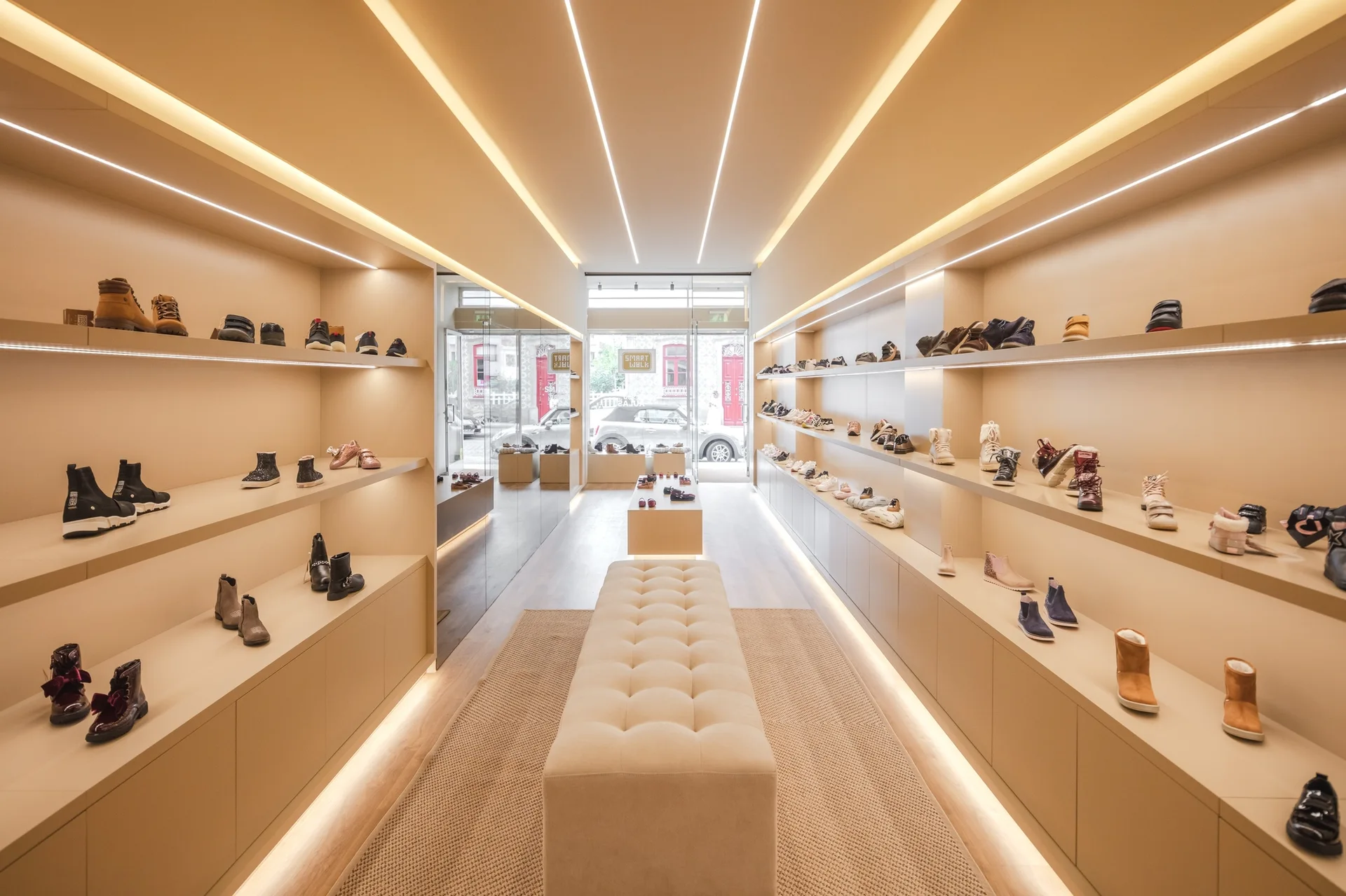A minimalist retail shoe store features warm beige shelves lined with shoes, a central cream tufted bench on a woven rug, and recessed linear lighting.