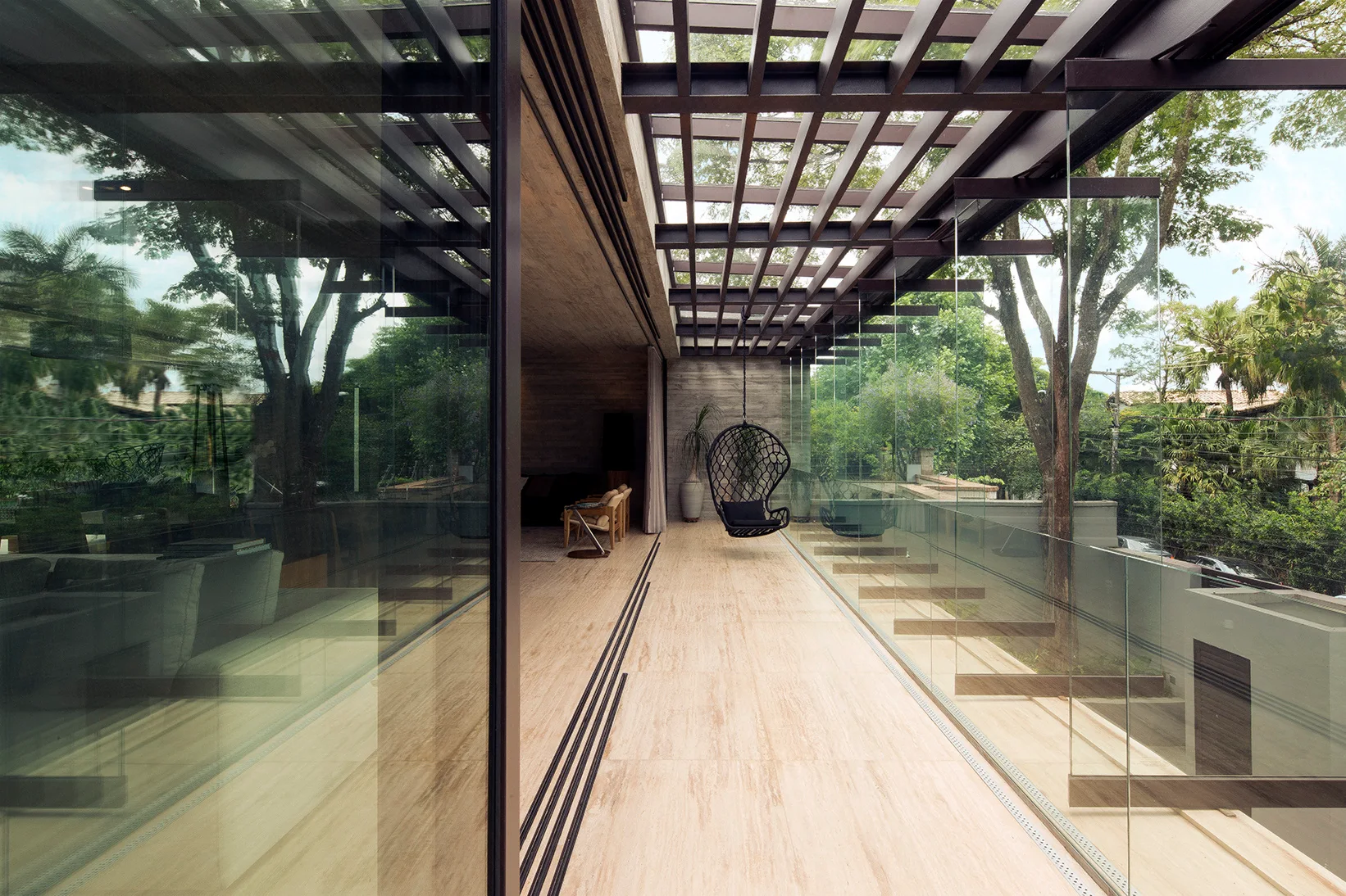 A modern concrete terrace features light beige travertine flooring, a black hanging chair, a dark metal structural pergola, and frameless glass railings.
