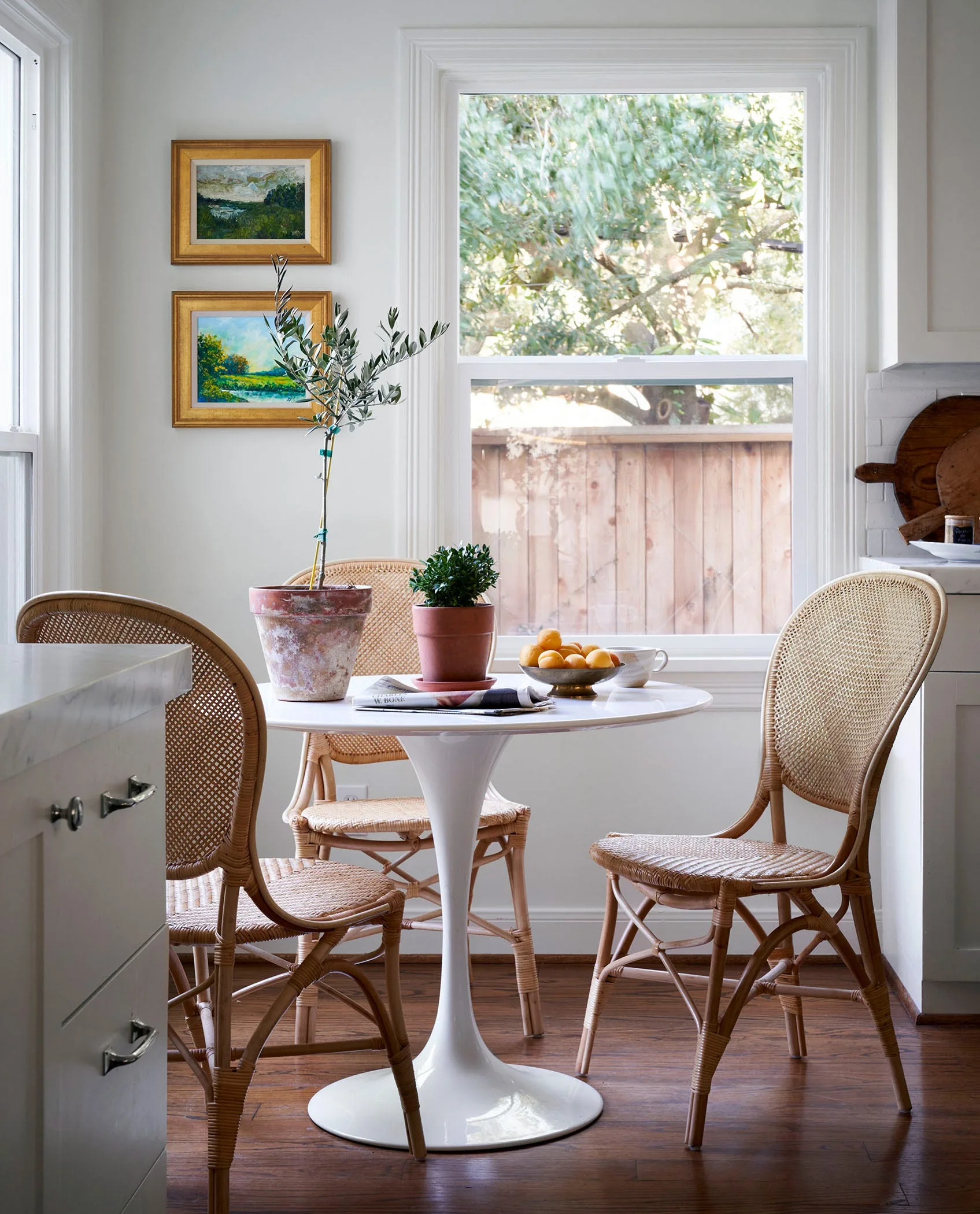 A sunlit kitchen breakfast nook features a round white pedestal table, three woven cane chairs, and two framed landscape paintings on the wall.