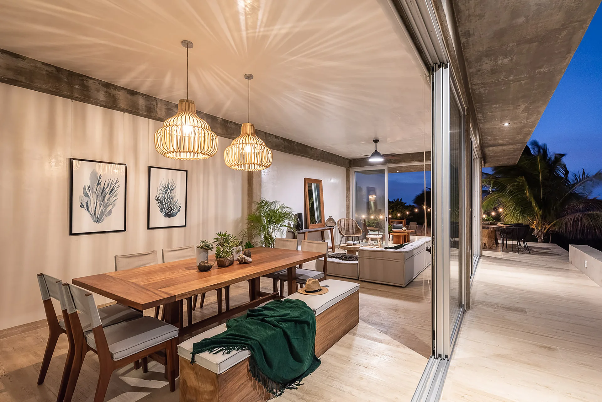 An open-concept dining room features a large wood table, upholstered bench, and two woven rattan pendant lights reflecting pattern on the ceiling.
