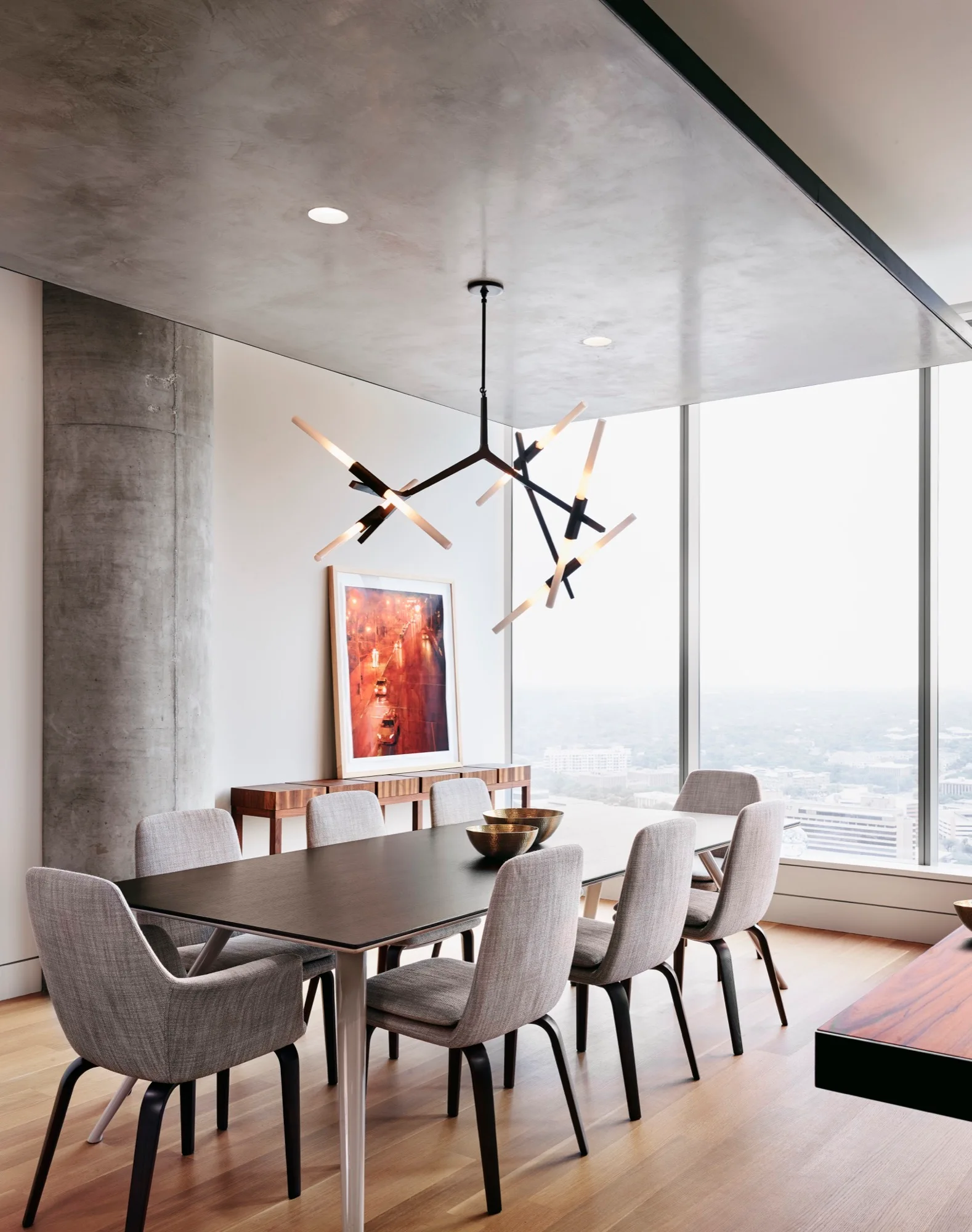 A modern dining room features a dark wood table with grey chairs, a branching black chandelier, and an exposed concrete column next to a large window.