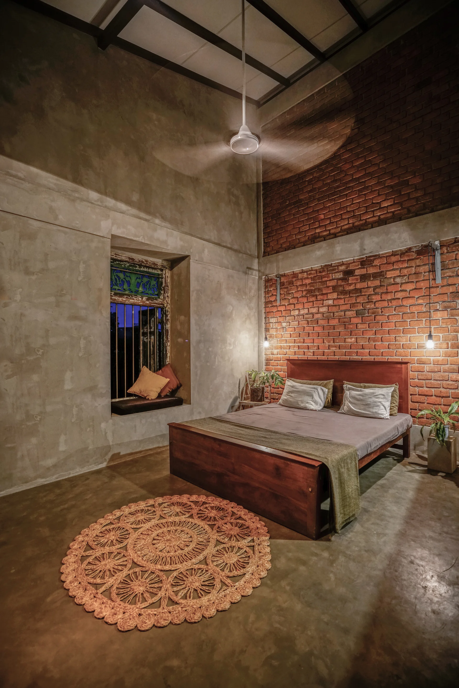 A rustic bedroom features a dark wooden bed against an exposed brick wall, a round woven rug on a polished concrete floor, and concrete walls.