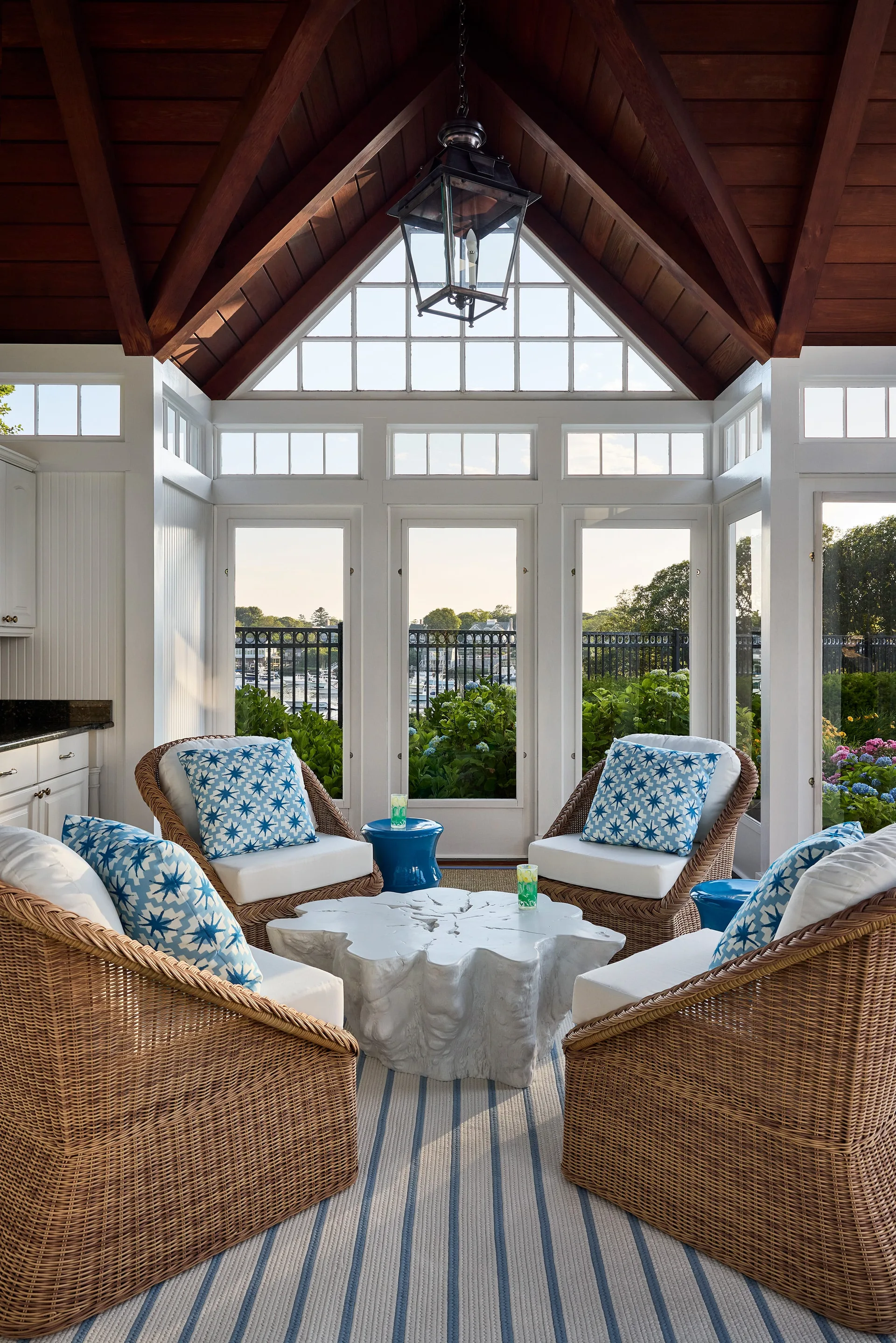 Four wicker armchairs with blue and white patterned pillows surround a white sculptural coffee table in a sunroom with a vaulted wood ceiling.