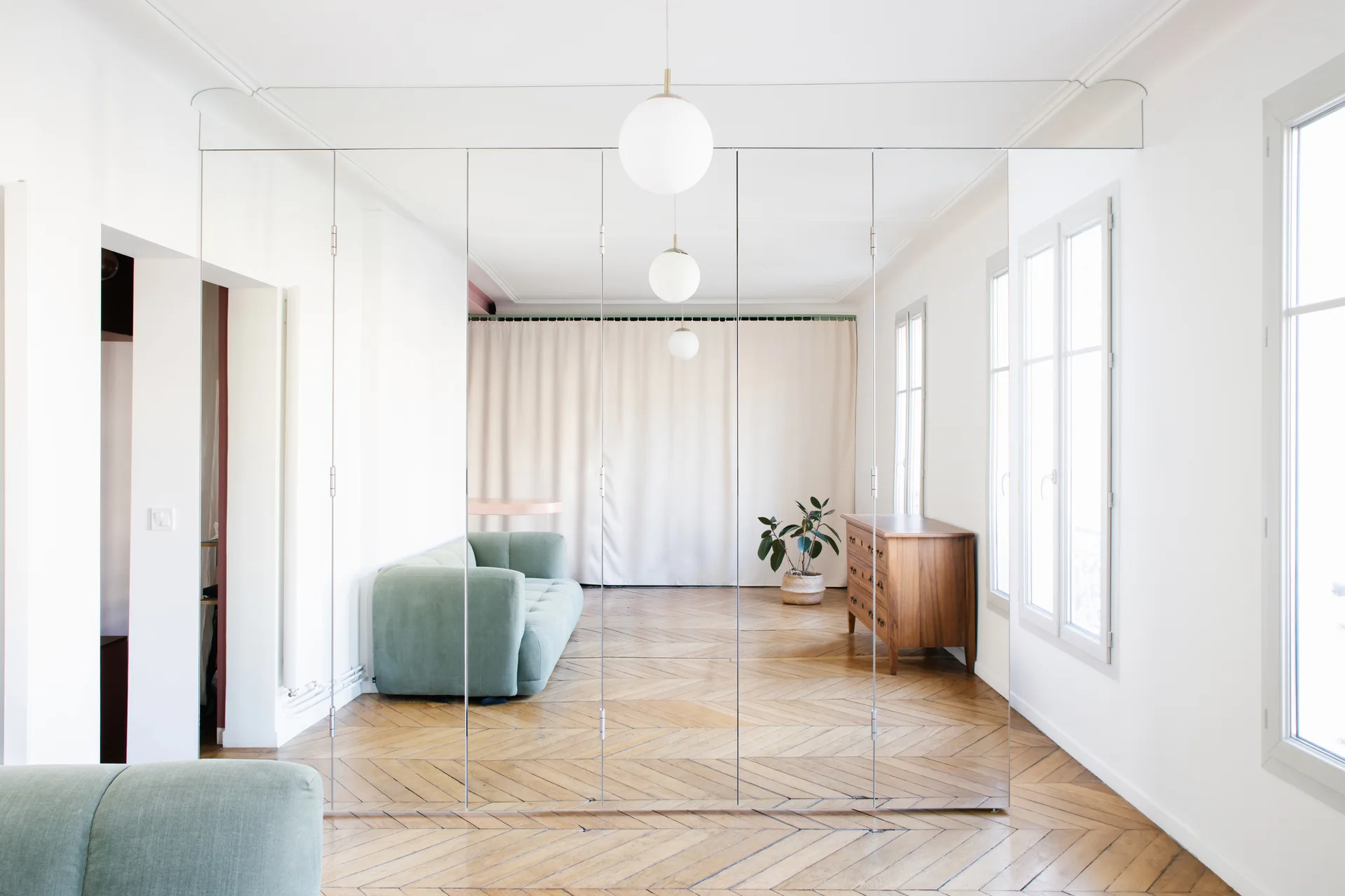 A wall of floor-to-ceiling mirror panels reflects a green sofa, a wooden dresser, and chevron parquet wood flooring in a bright white room.