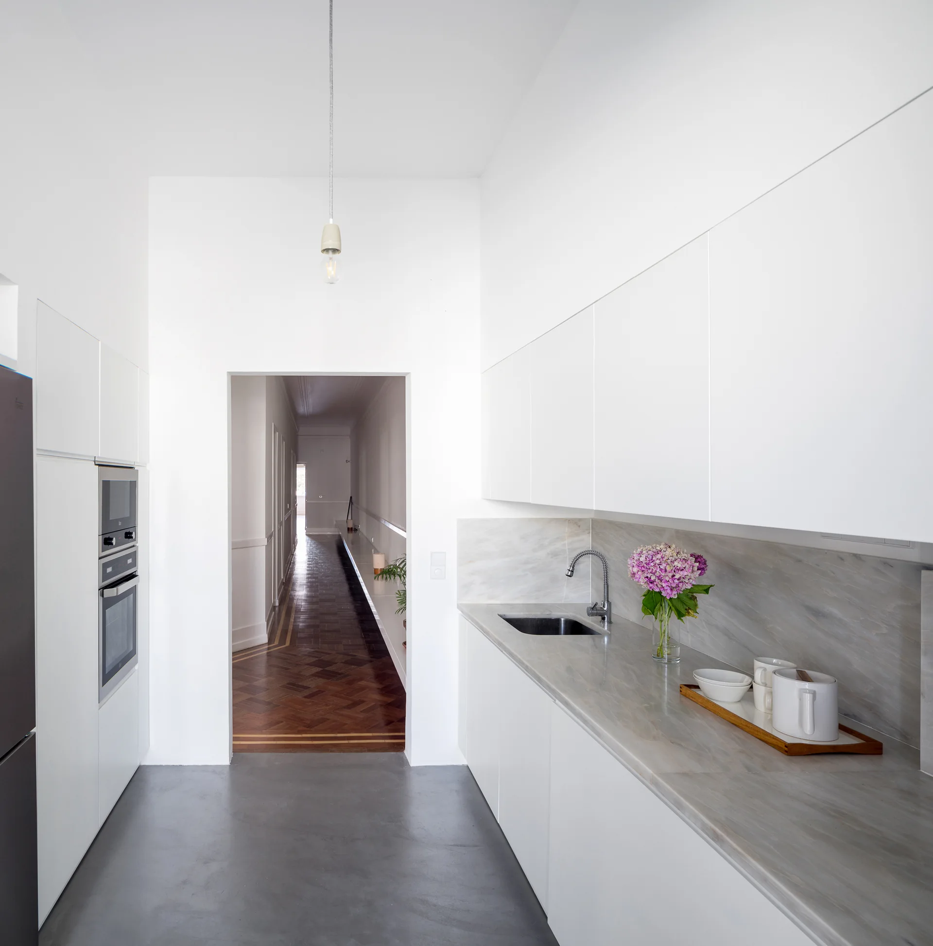 A minimalist white kitchen features flat-panel cabinets, a veined stone countertop, grey floors, and a view into a long wood herringbone hallway.