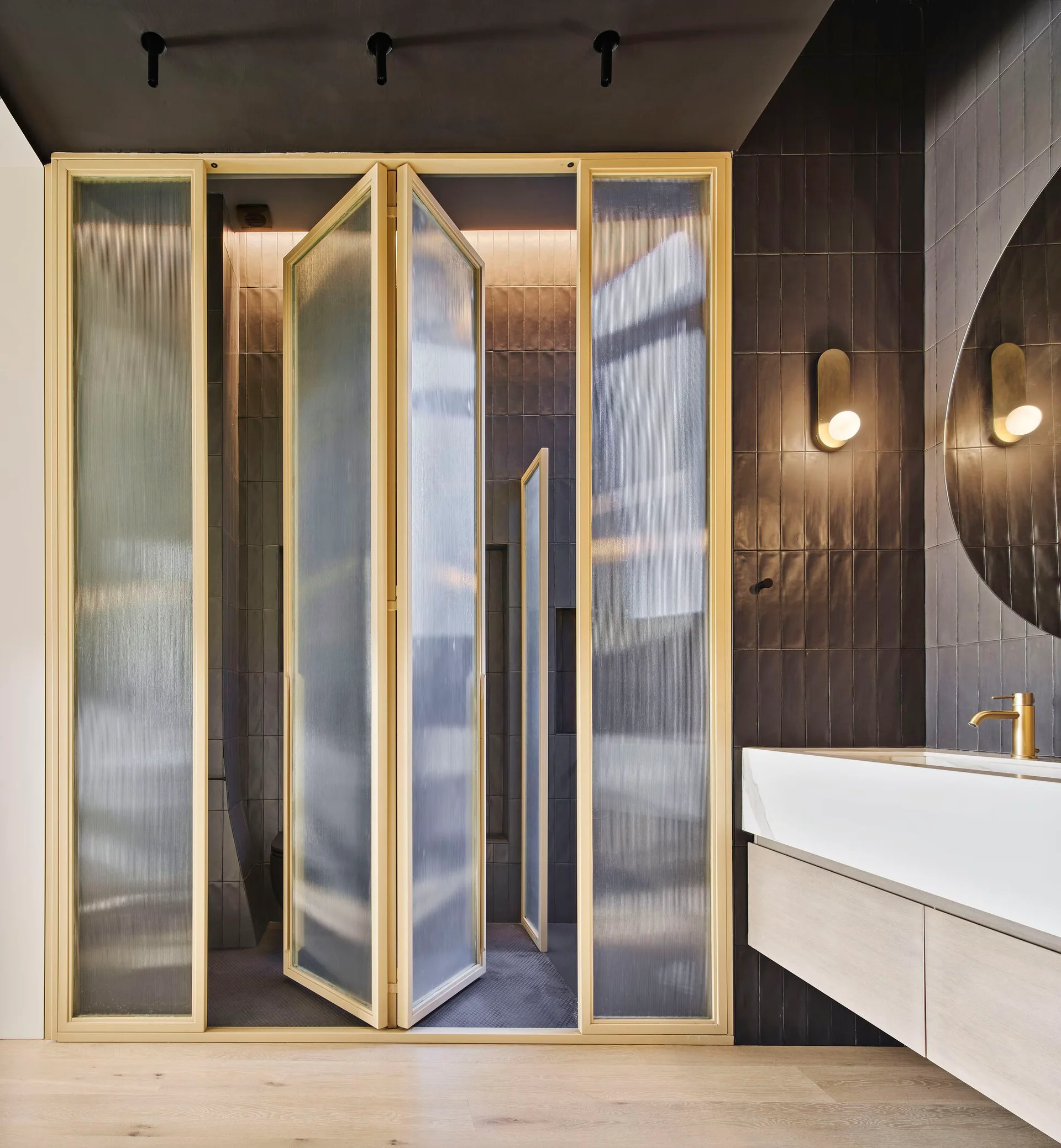 A modern bathroom interior with a fluted glass folding door partition, dark tiled walls, and a floating wood vanity with gold fixtures.