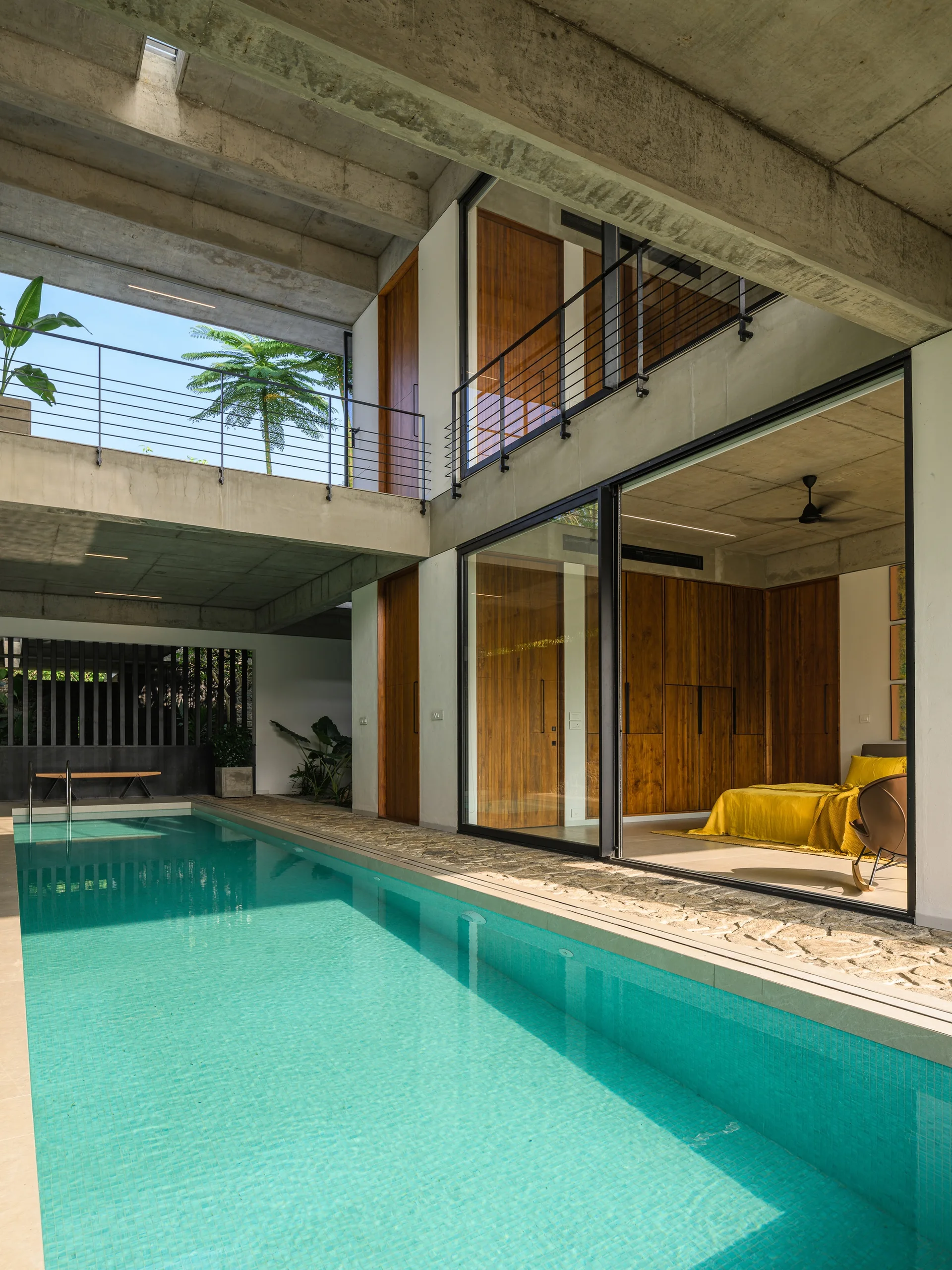 A modern home features a rectangular turquoise lap pool beneath exposed concrete beams, adjacent to a bedroom with sliding glass doors.