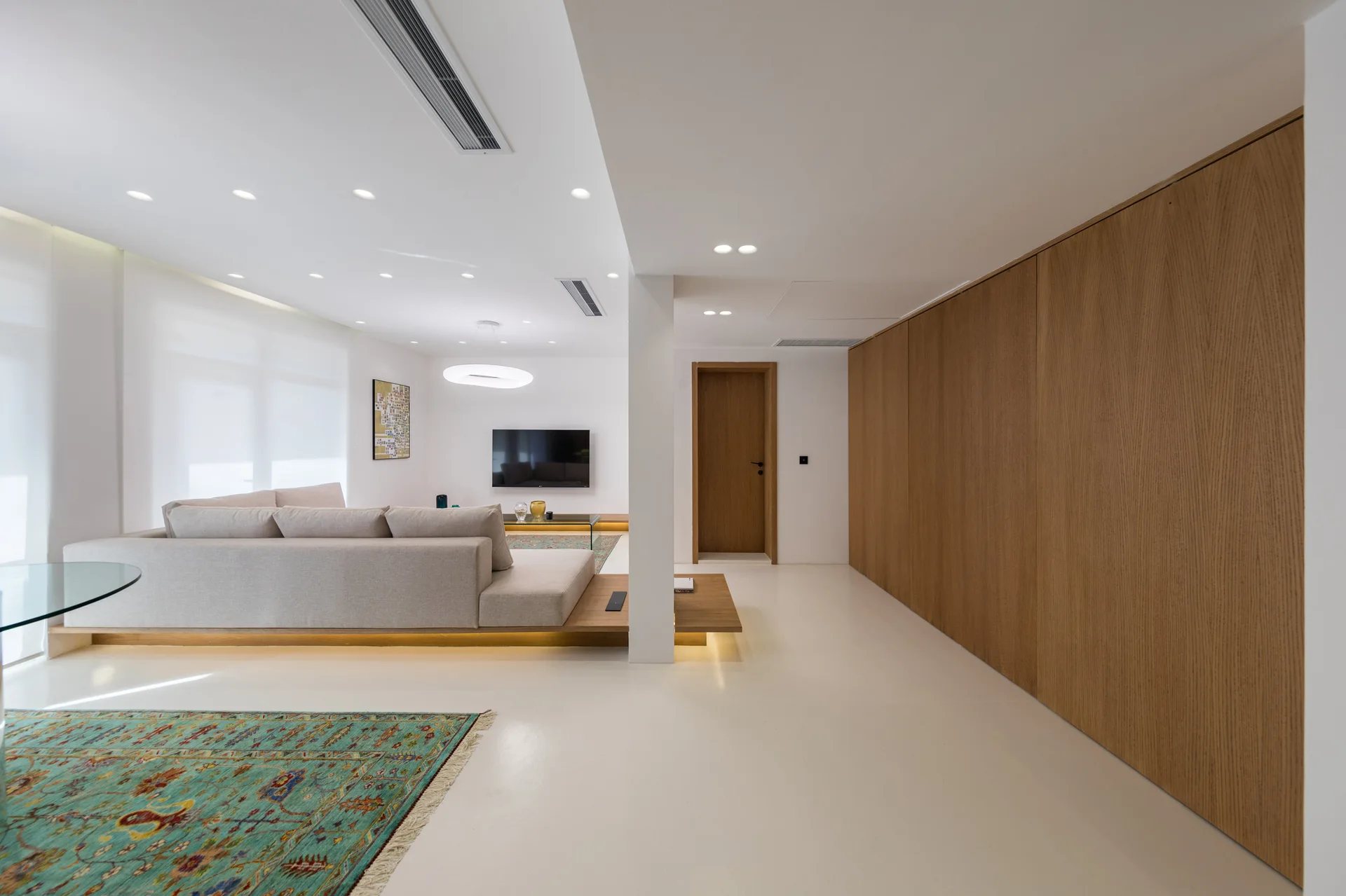 A beige modular sectional sits on a low-profile wood platform with integrated lighting next to a full-height oak wood paneled storage wall.