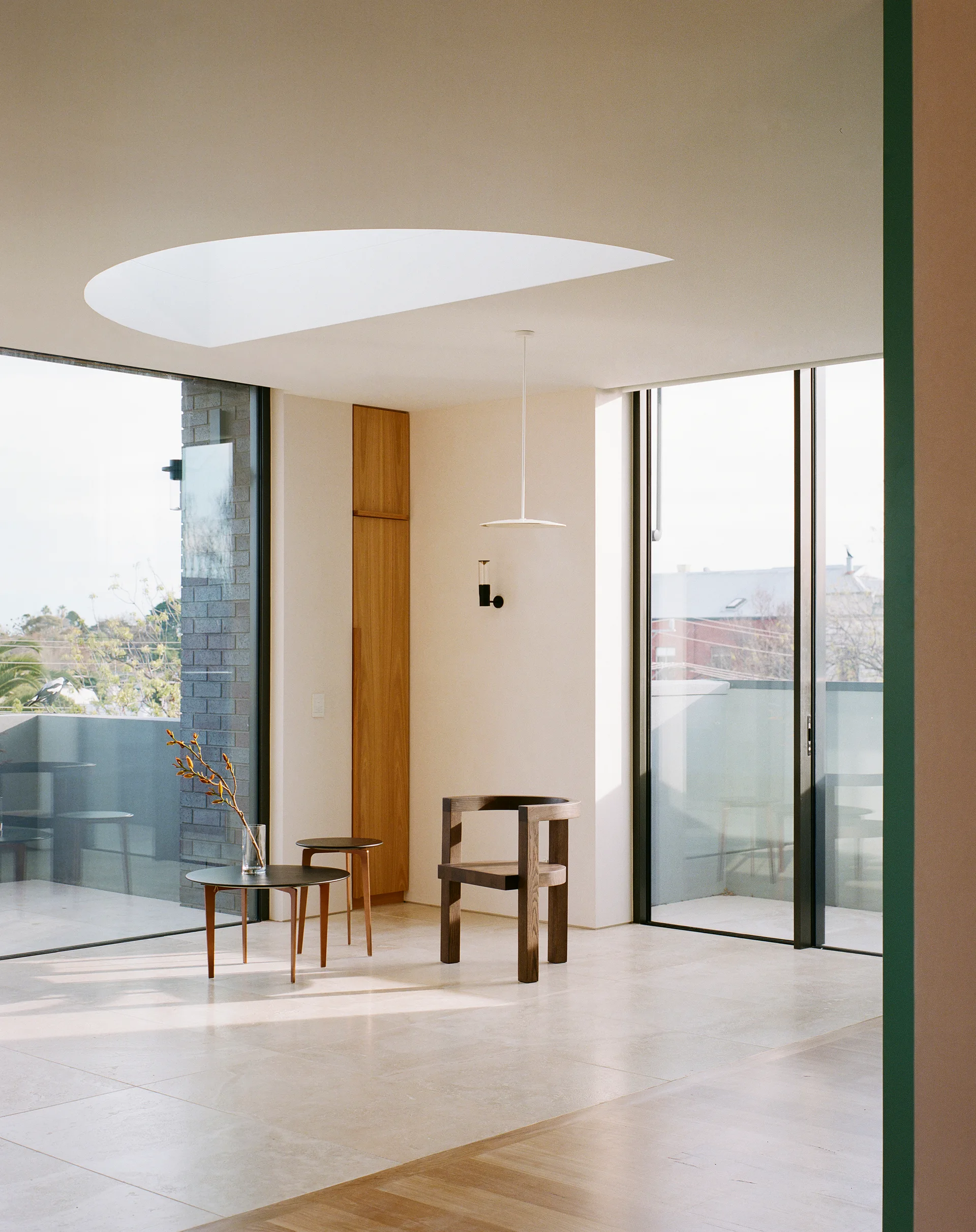 A minimalist room with stone tile flooring features a sculptural wood armchair, nesting side tables, and a curved skylight near large glass doors.