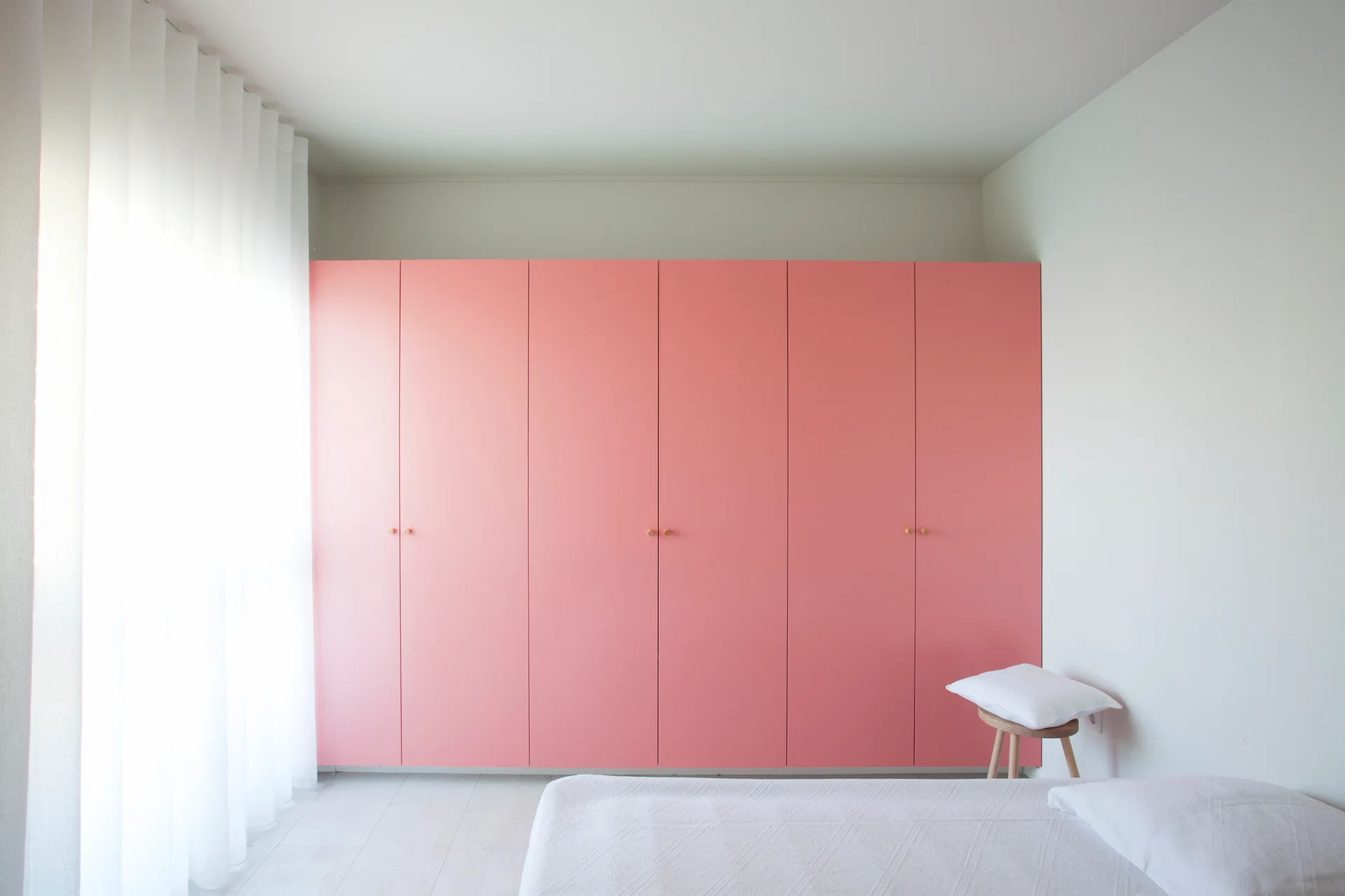 A minimalist bedroom features a large, matte pink built-in wardrobe next to a window with sheer white curtains and a low bed with white bedding.