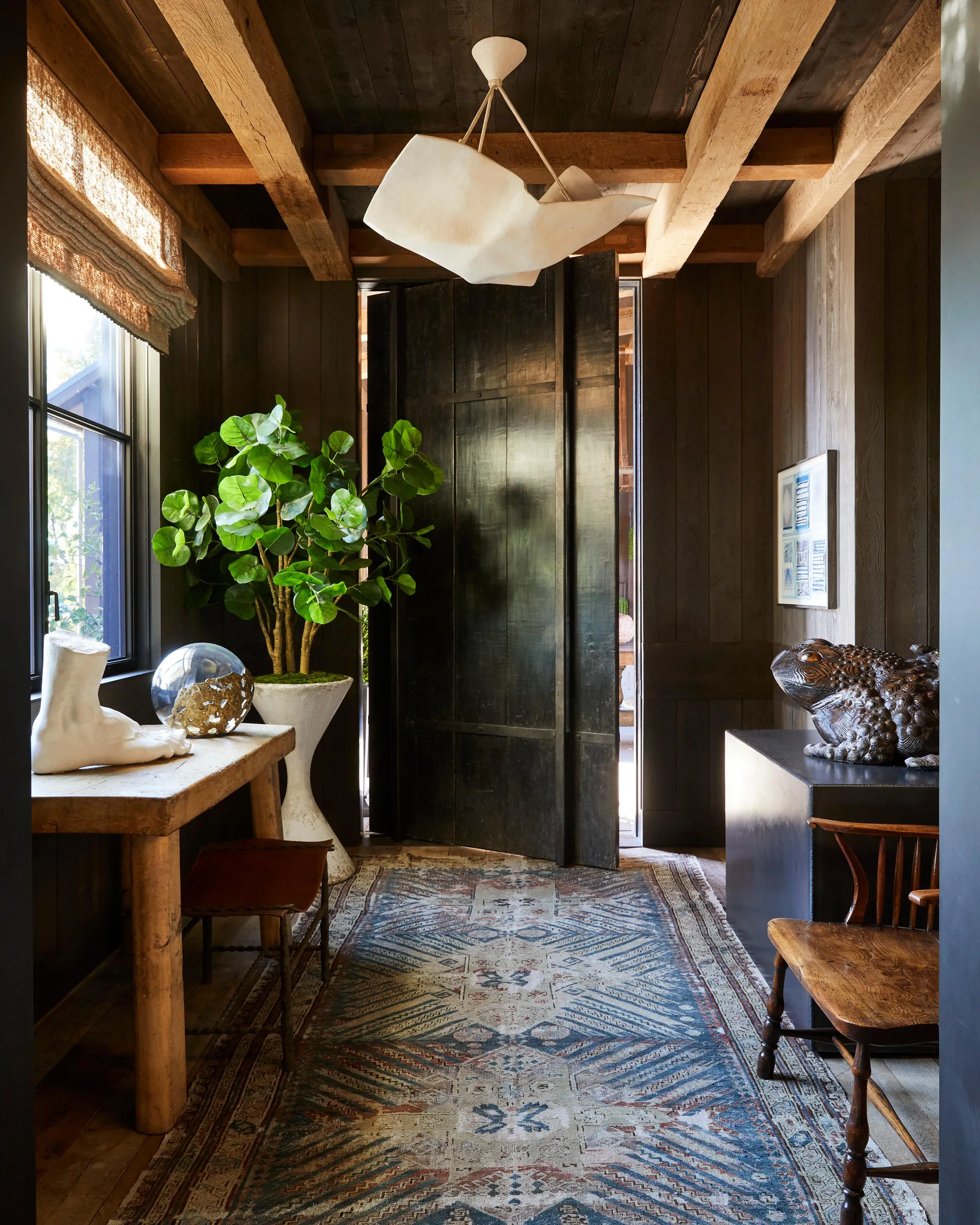 A rustic entryway features dark wood paneled walls, heavy ceiling beams, a patterned blue area rug, and a large green plant in a white pedestal.
