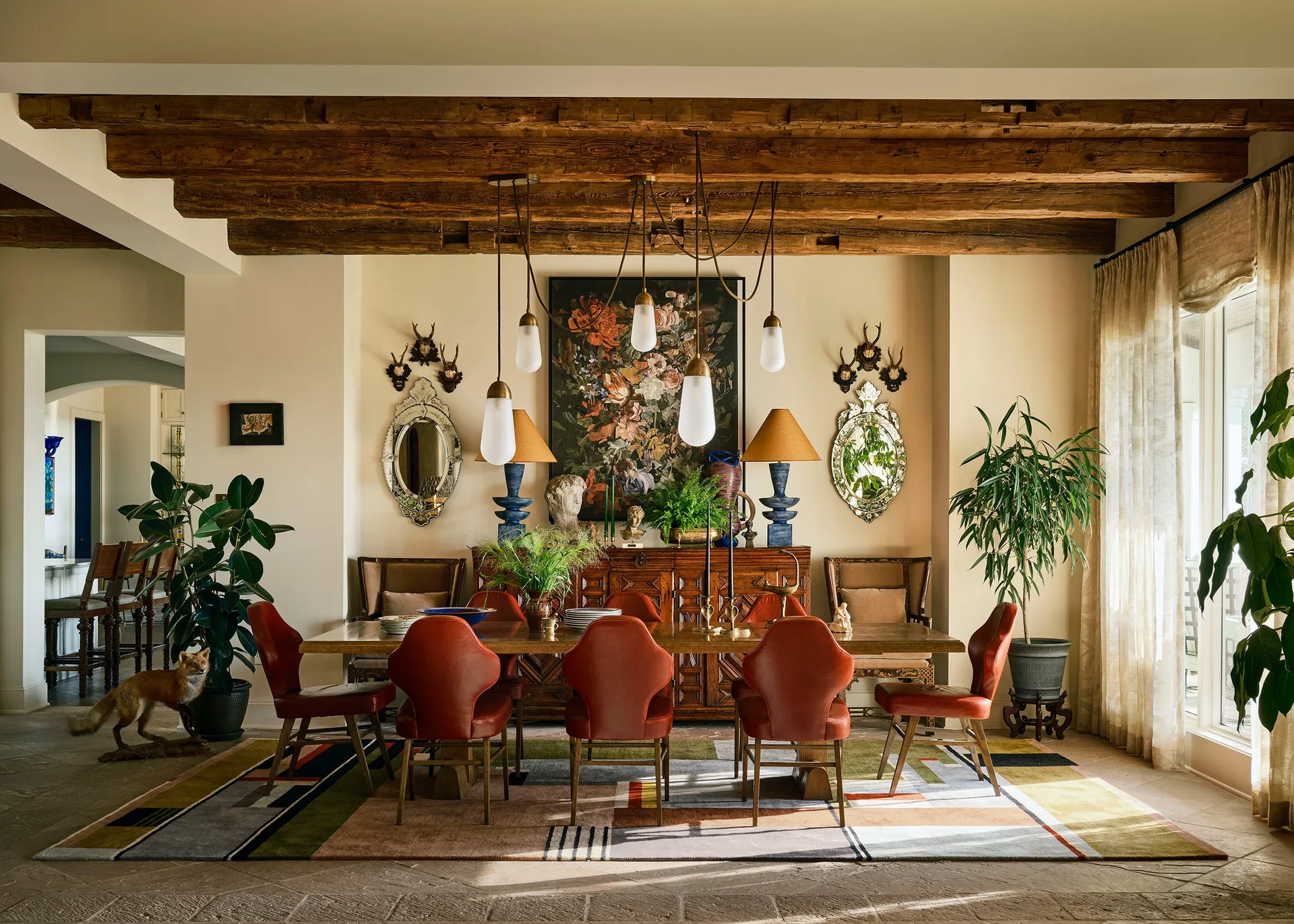 An eclectic dining room features red leather chairs around a wooden table beneath exposed timber beams and a modern multi-pendant light fixture.