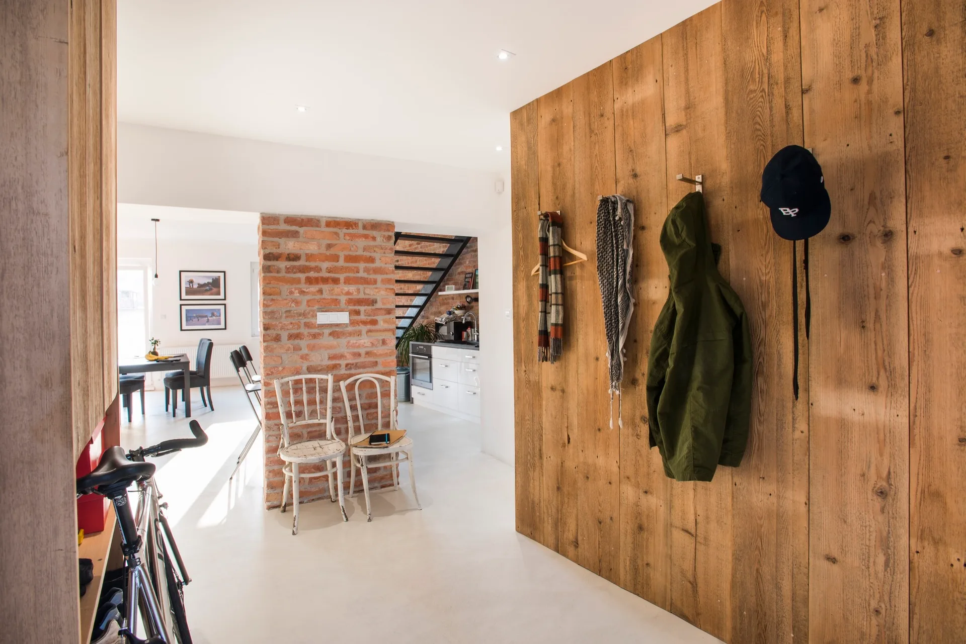An entryway features a natural wood plank wall with coat hooks, an exposed brick column with two white chairs, and a white matte floor.