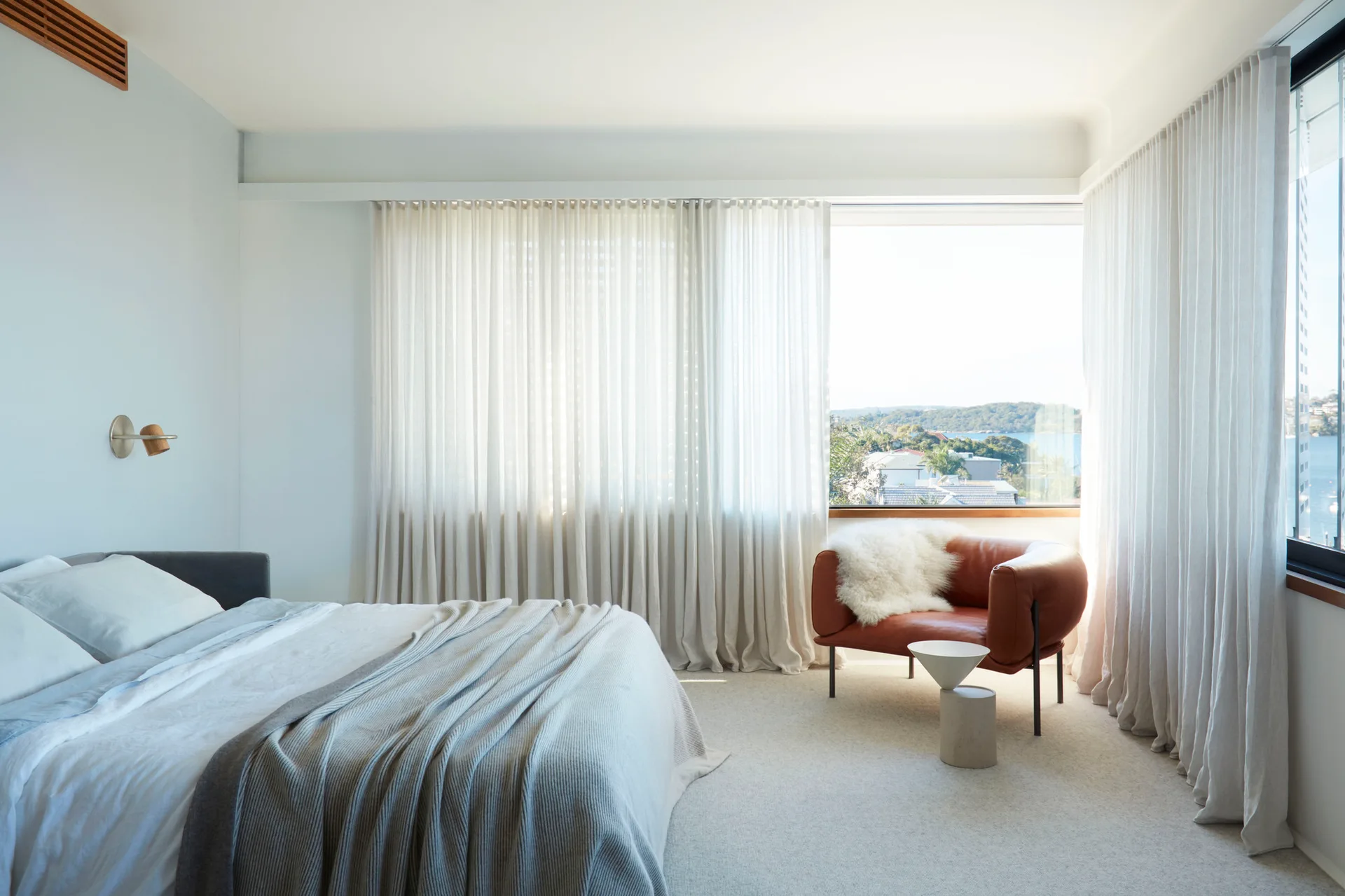 A minimalist bedroom features a bed with grey linen sheets, a tan leather armchair with sheepskin, and full-length sheer curtains.