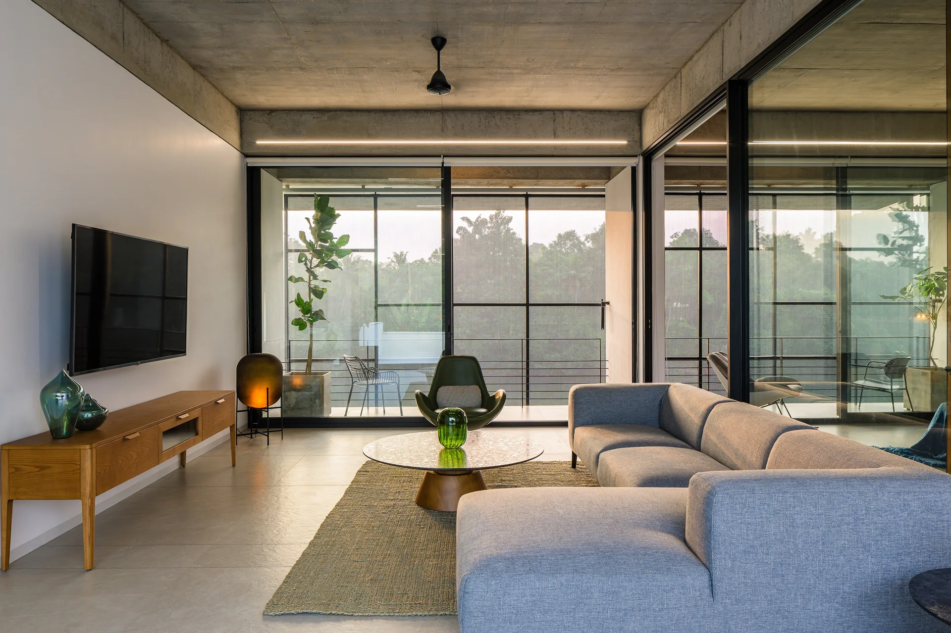 A modern living room features a grey sectional sofa, a low wooden media console, and an exposed concrete ceiling facing large sliding glass balcony doors.