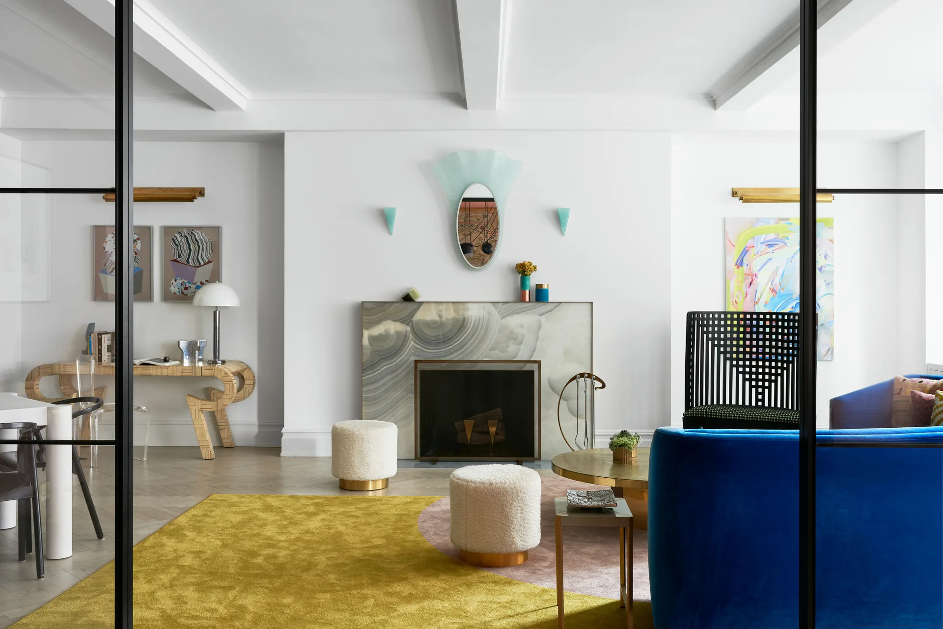 An eclectic living room seen through a black framed glass door, featuring a swirling stone fireplace, a curved blue sofa, and white boucle stools.