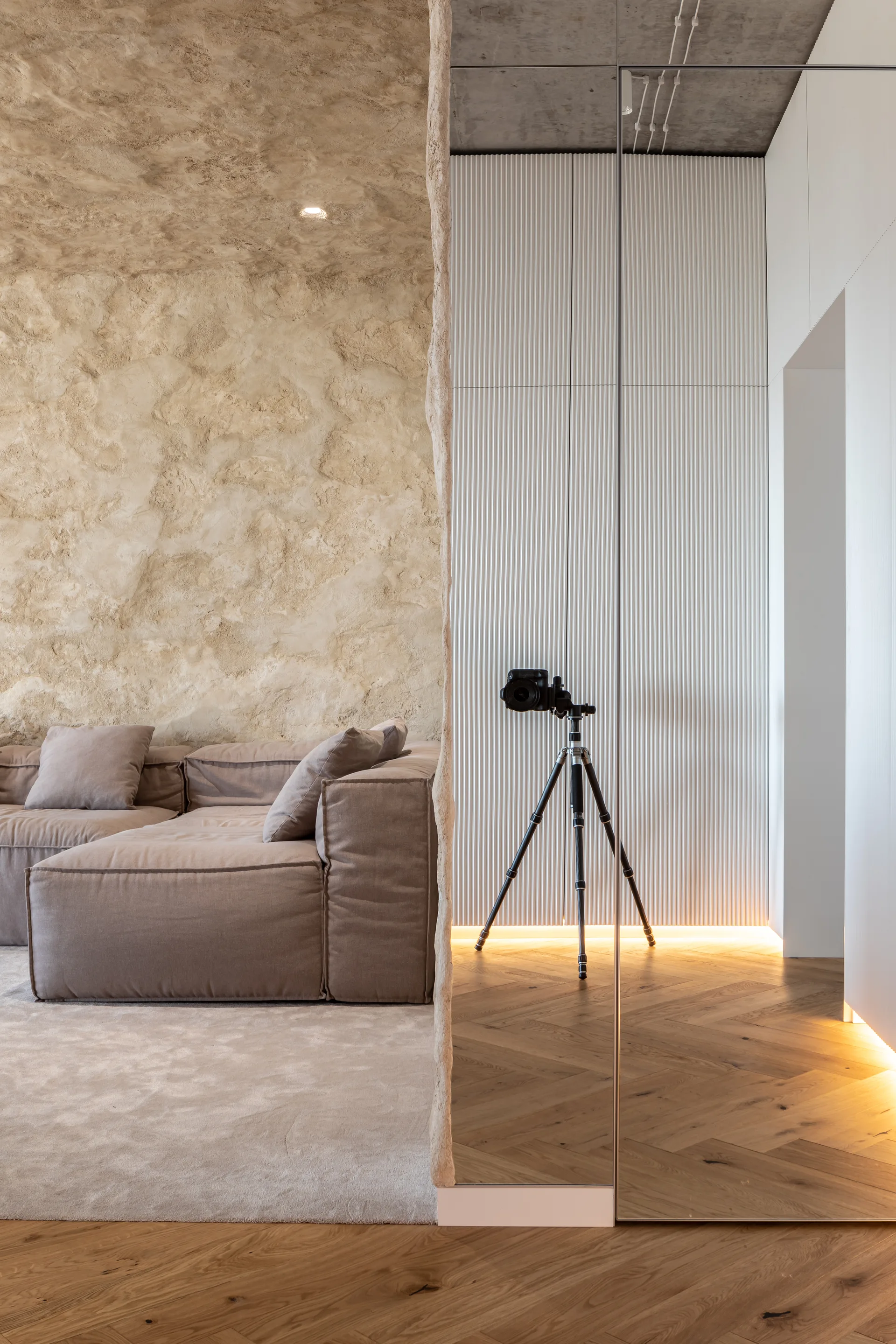 A modular beige sofa sits before a rough stone wall, adjacent to a mirrored wall, fluted white panels, and herringbone wood flooring.