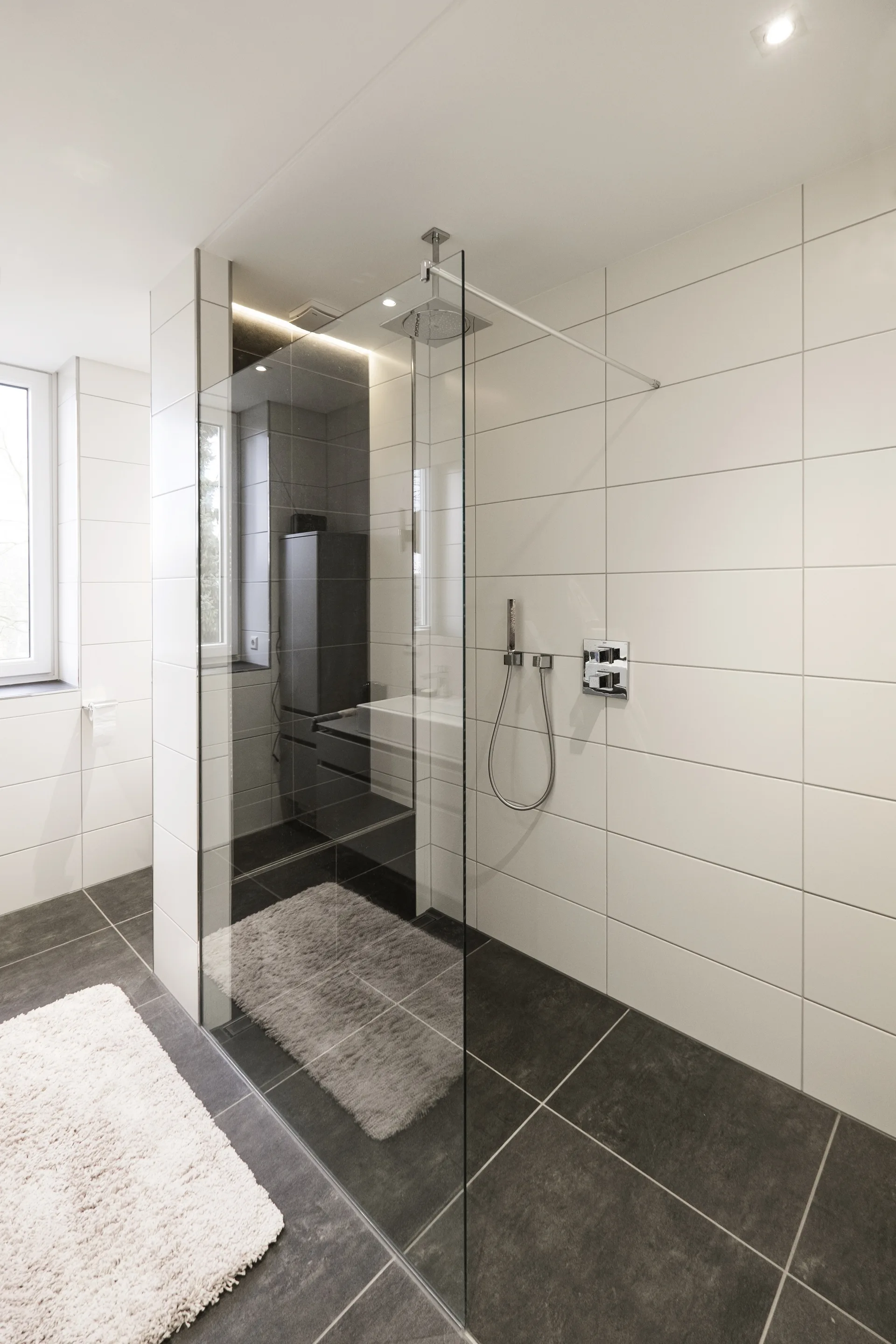 A modern walk-in shower features a single frameless glass partition, large white rectangular wall tiles, and large dark grey floor tiles.
