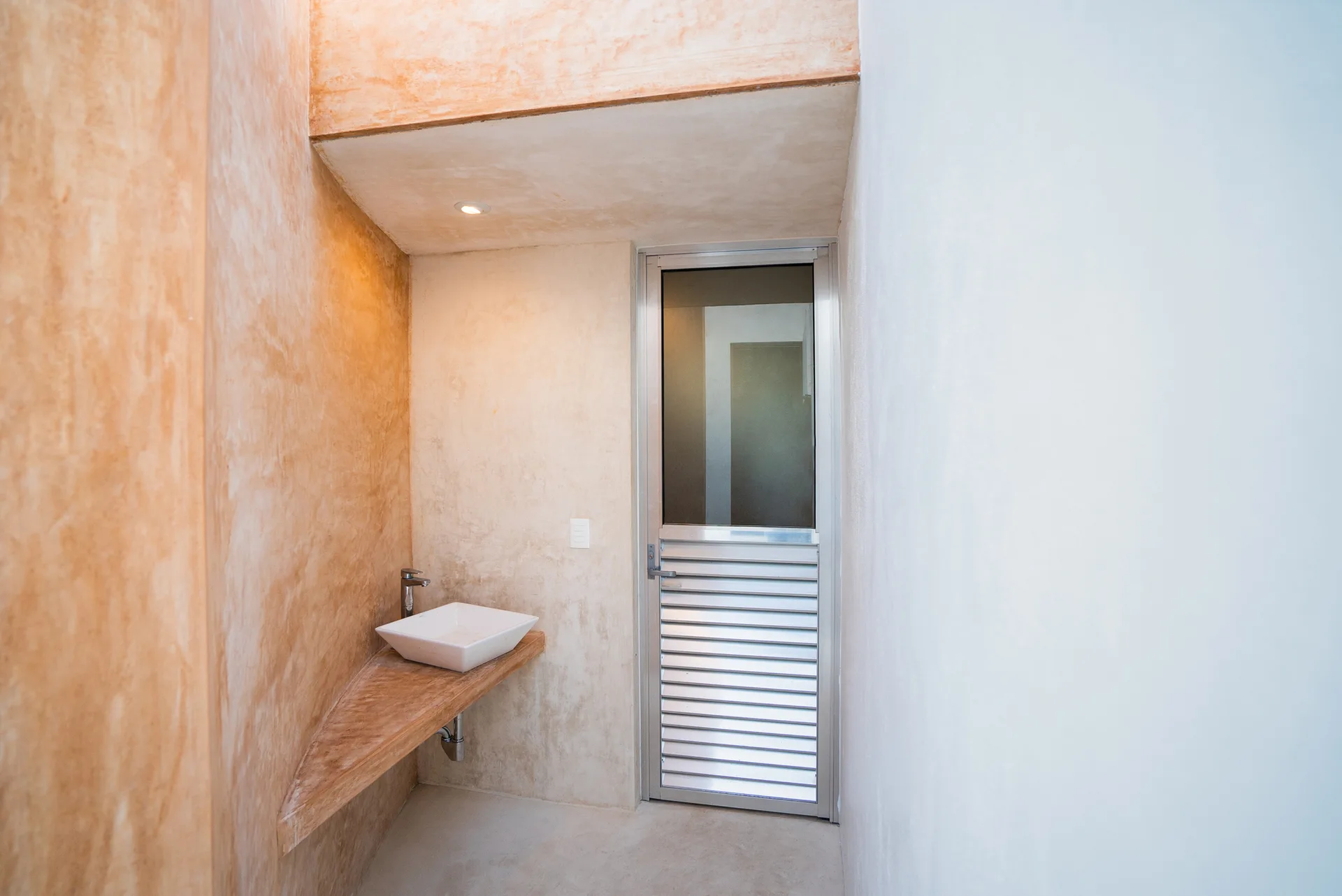 A minimalist bathroom alcove features a white vessel sink on a triangular wood vanity shelf against textured plaster walls next to a glass door.